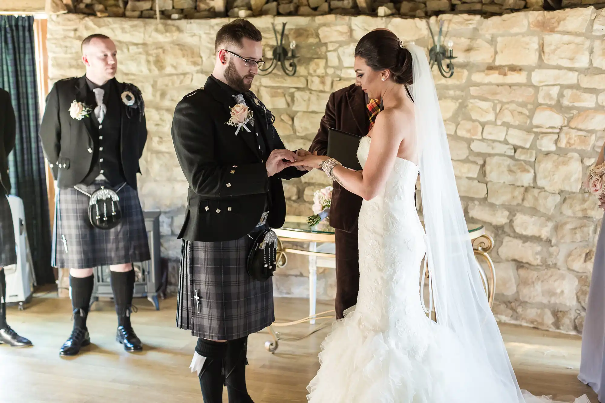 A groom in a black jacket and tartan kilt places a ring on the bride’s finger during their wedding ceremony. The bride wears a strapless white gown and long veil. A best man in a kilt stands nearby. They are indoors with a stone wall background.