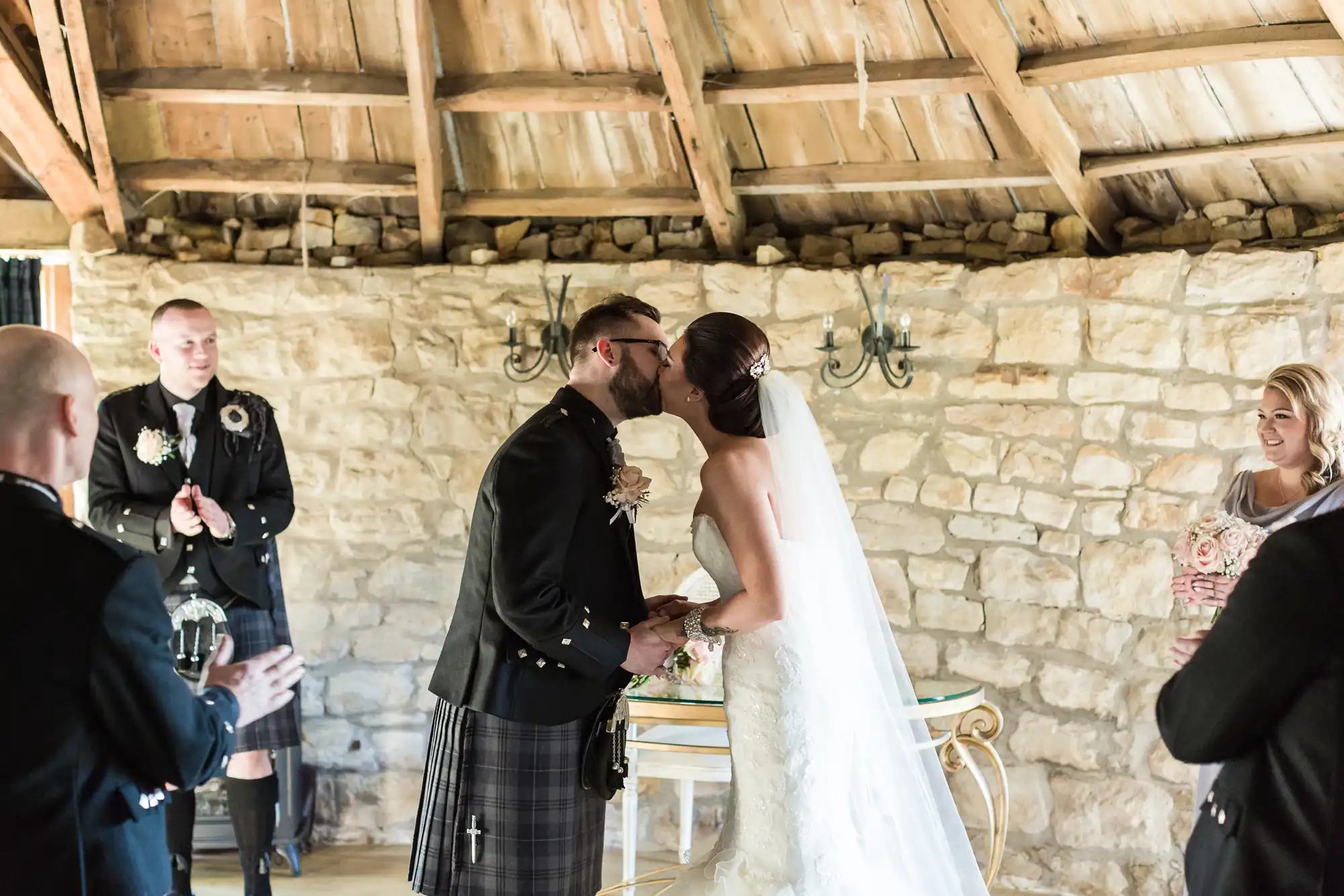A bride in a white dress and a groom in a kilt kiss at their wedding ceremony inside a rustic stone building. Bridesmaid and groomsmen stand nearby, smiling and applauding. The wooden ceiling and pale stone walls create a cosy, intimate atmosphere.