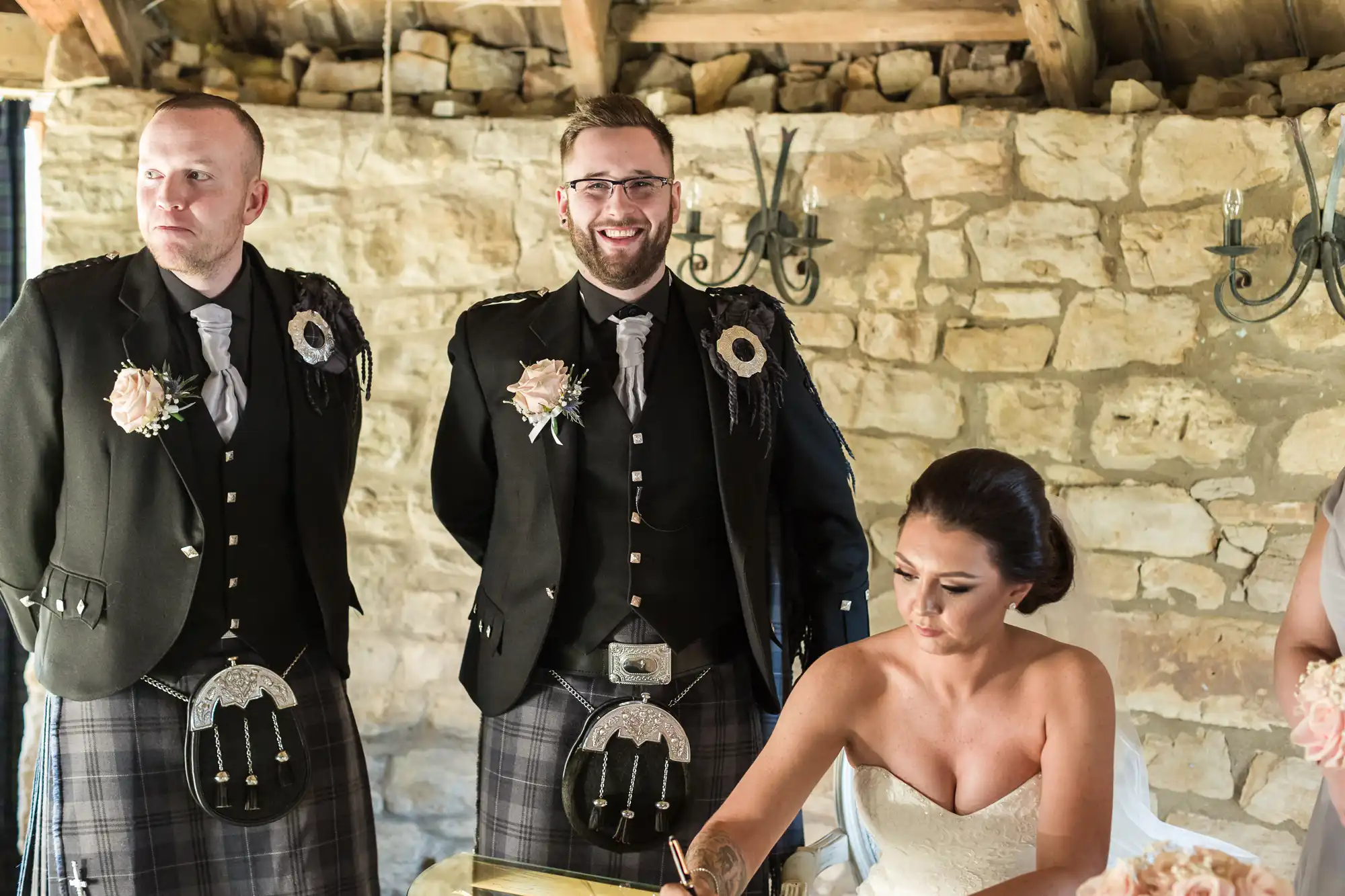 A bride in a strapless white dress signs a document at a table. Two men in black jackets, kilts, and buttonholes stand behind her, one smiling. They are in a rustic stone-walled room with exposed beams and wall lights.