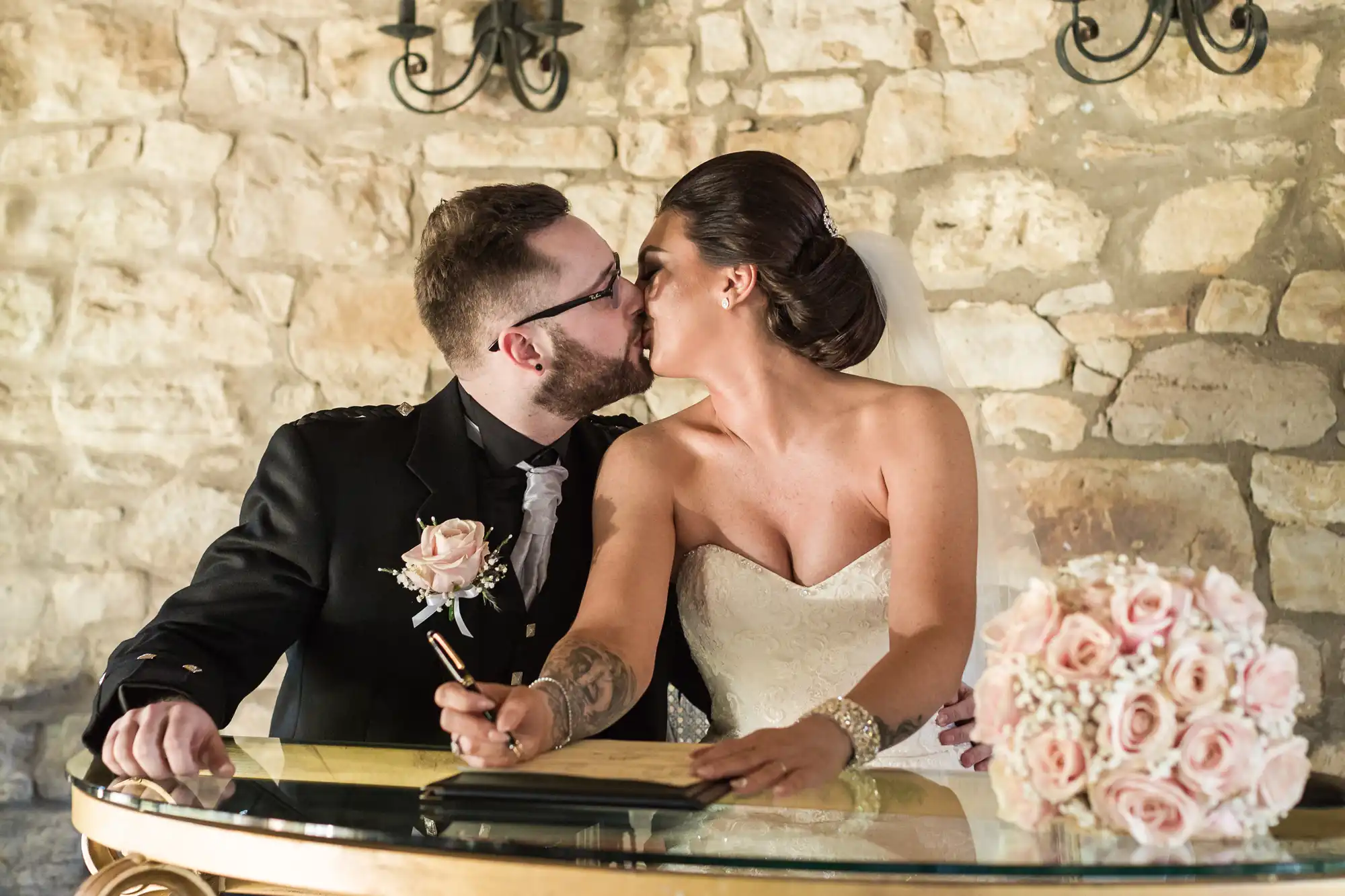 A bride in a strapless white dress and veil kisses a groom in a dark suit at a glass table. She holds a pen and bouquet of pink roses. Stone wall background, soft lighting, wedding atmosphere.