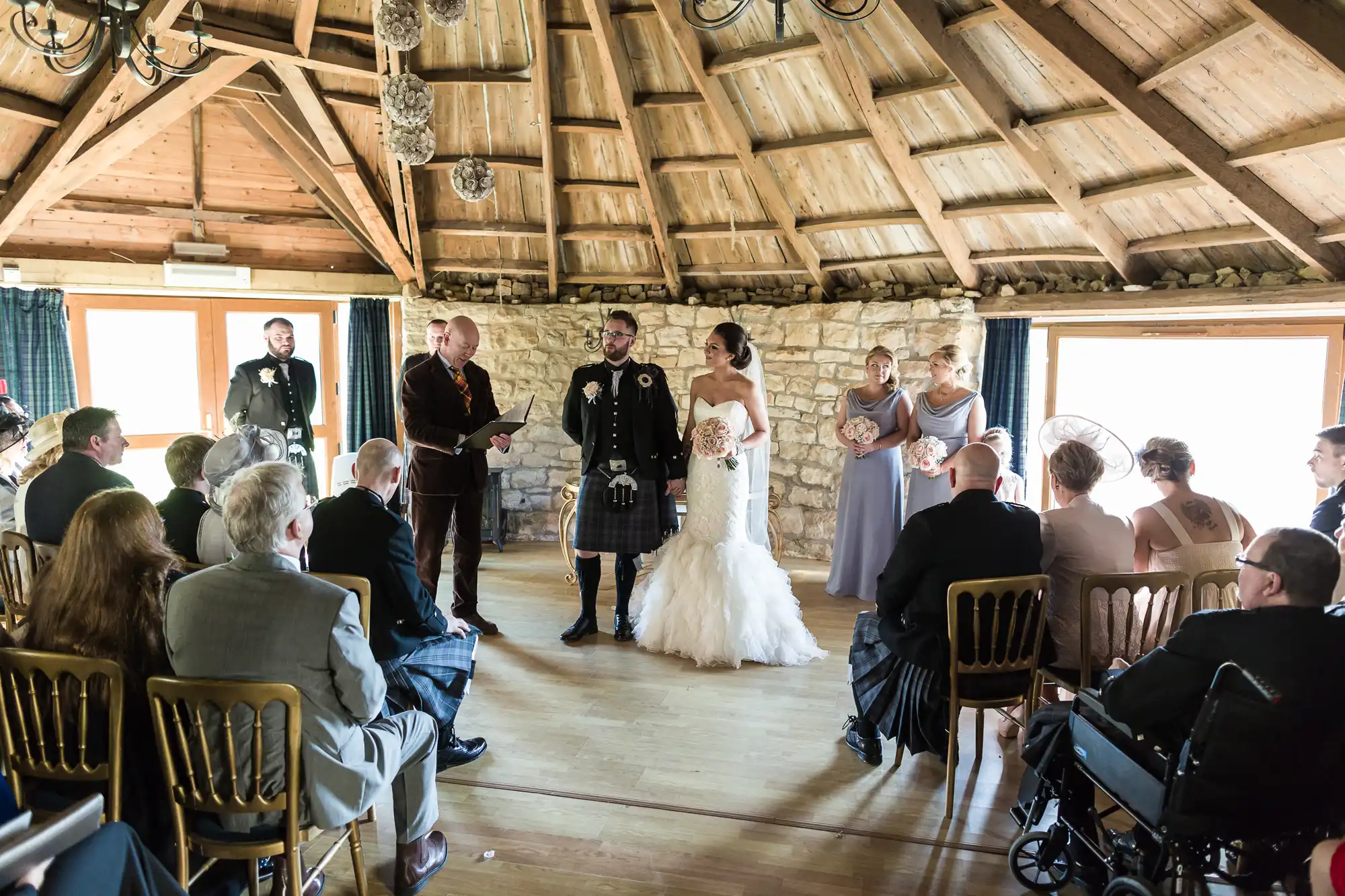 A bride in a white dress and groom in a kilt stand at the altar in a rustic, wood-beamed room with stone walls. Bridesmaids in lavender dresses, guests seated on gold chairs, and a man in a wheelchair are visible, as sunlight streams in through large windows.