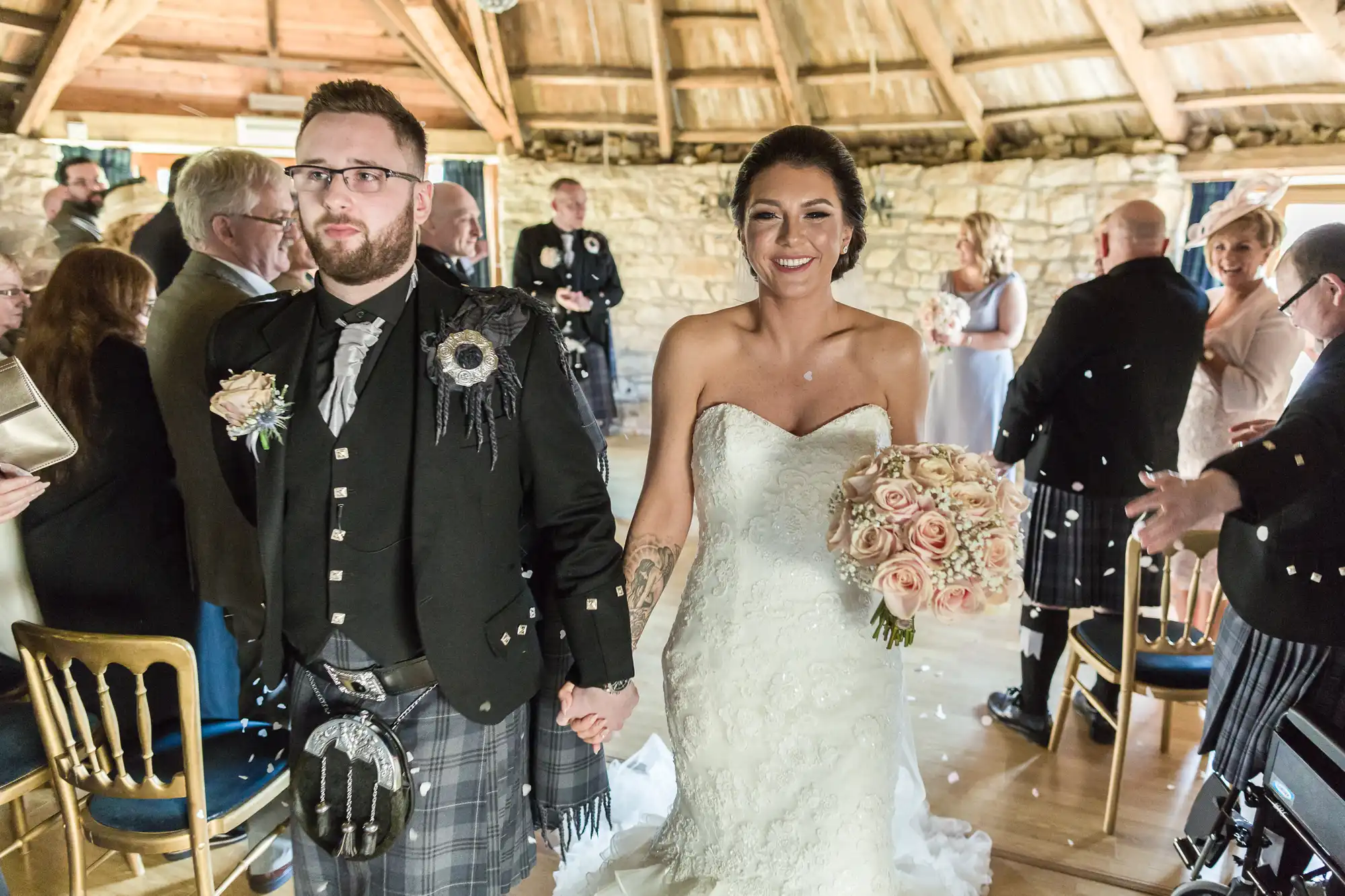 A bride in a strapless white dress holds a bouquet of pink roses, smiling and walking down the aisle beside a groom in a black jacket and tartan kilt. Guests stand and applaud inside a rustic venue with wooden beams and stone walls.