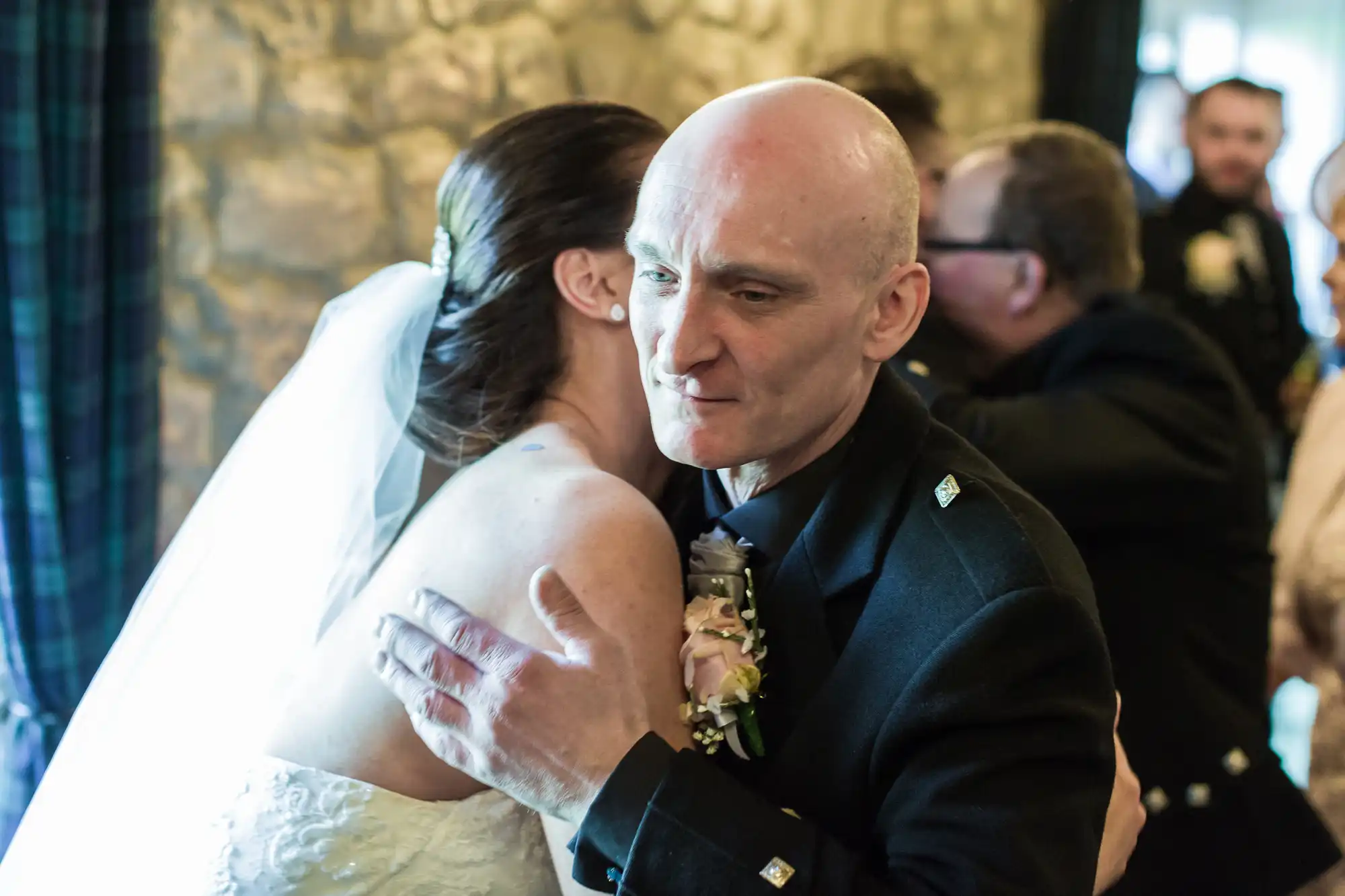 A bride in a white dress and veil hugs a man in a black suit with a buttonhole. The man looks emotional, eyes slightly closed. They are indoors, near a stone wall, with several blurred people in the background.