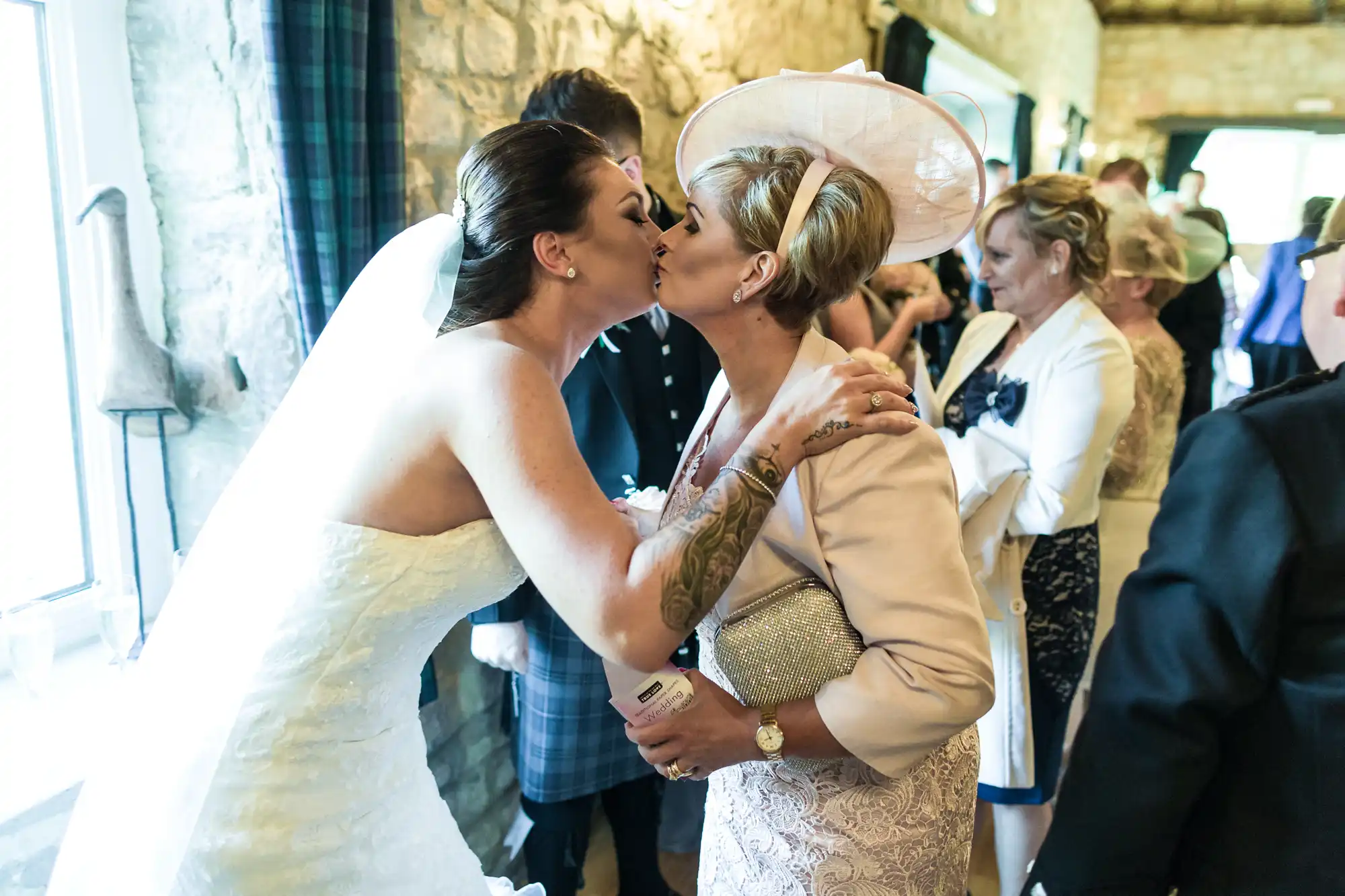 A bride in a white strapless gown and veil shares a kiss on the lips with an older woman wearing a pale pink outfit and matching hat at a wedding reception, surrounded by other well-dressed guests in a rustic stone-walled venue.