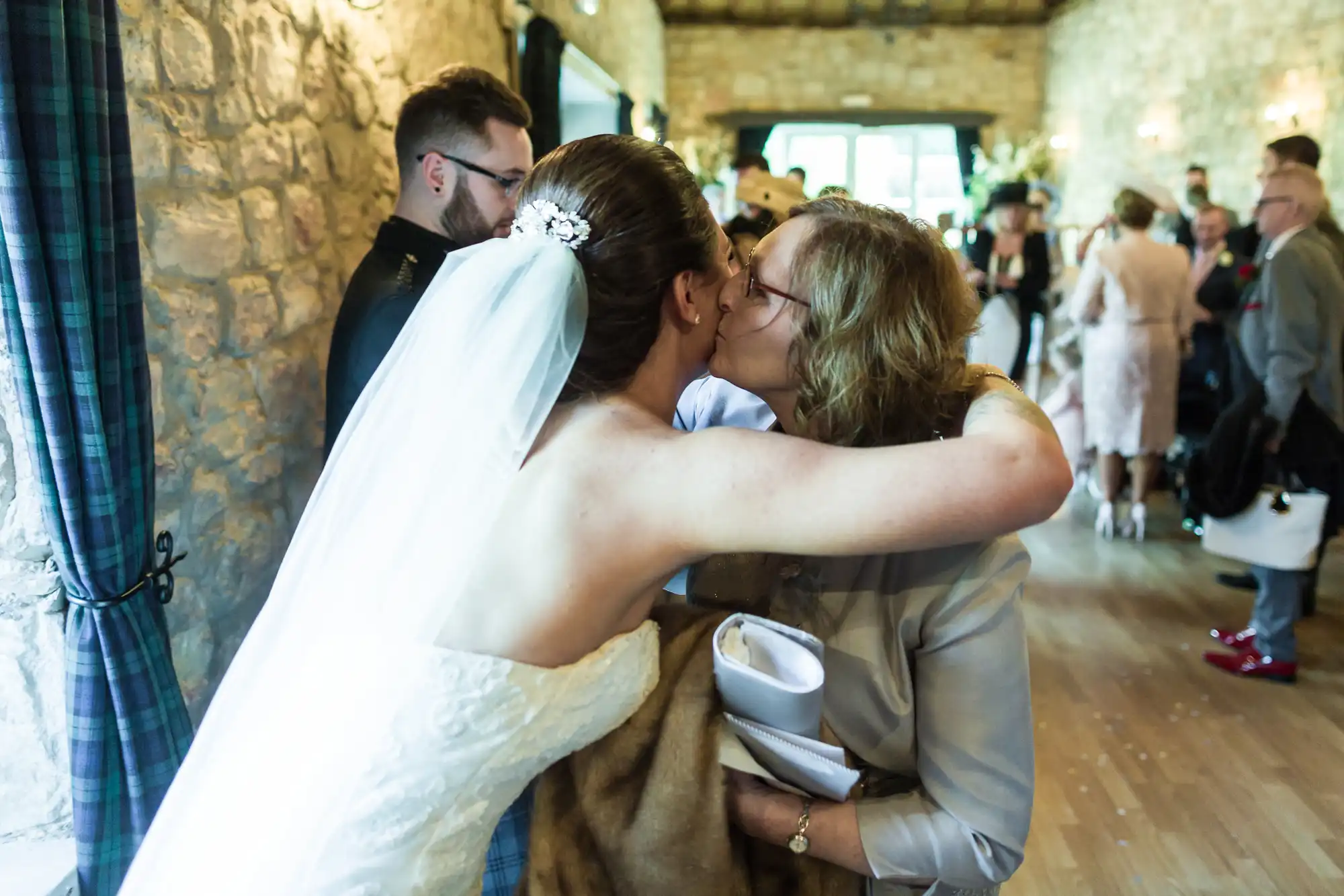 A bride in a white dress and veil hugs and kisses a woman on the cheek in a warmly lit stone hall, surrounded by guests in formal attire blurred in the background.