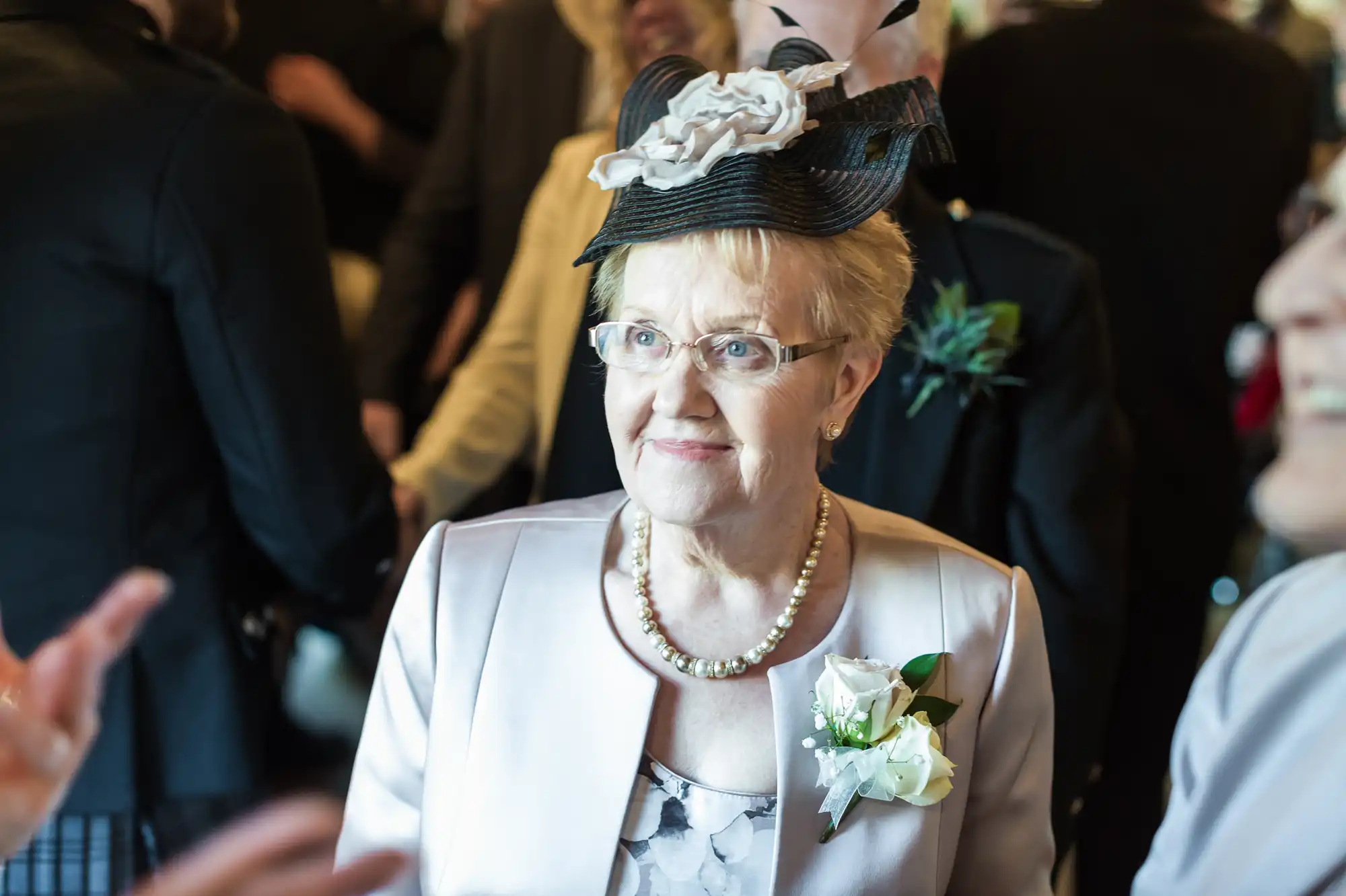 An elderly woman with short blonde hair, glasses, and a decorative black and white hat smiles. She wears a light jacket, pearl necklace, and a white rose corsage. People in formal attire are blurred in the background, suggesting a special event.