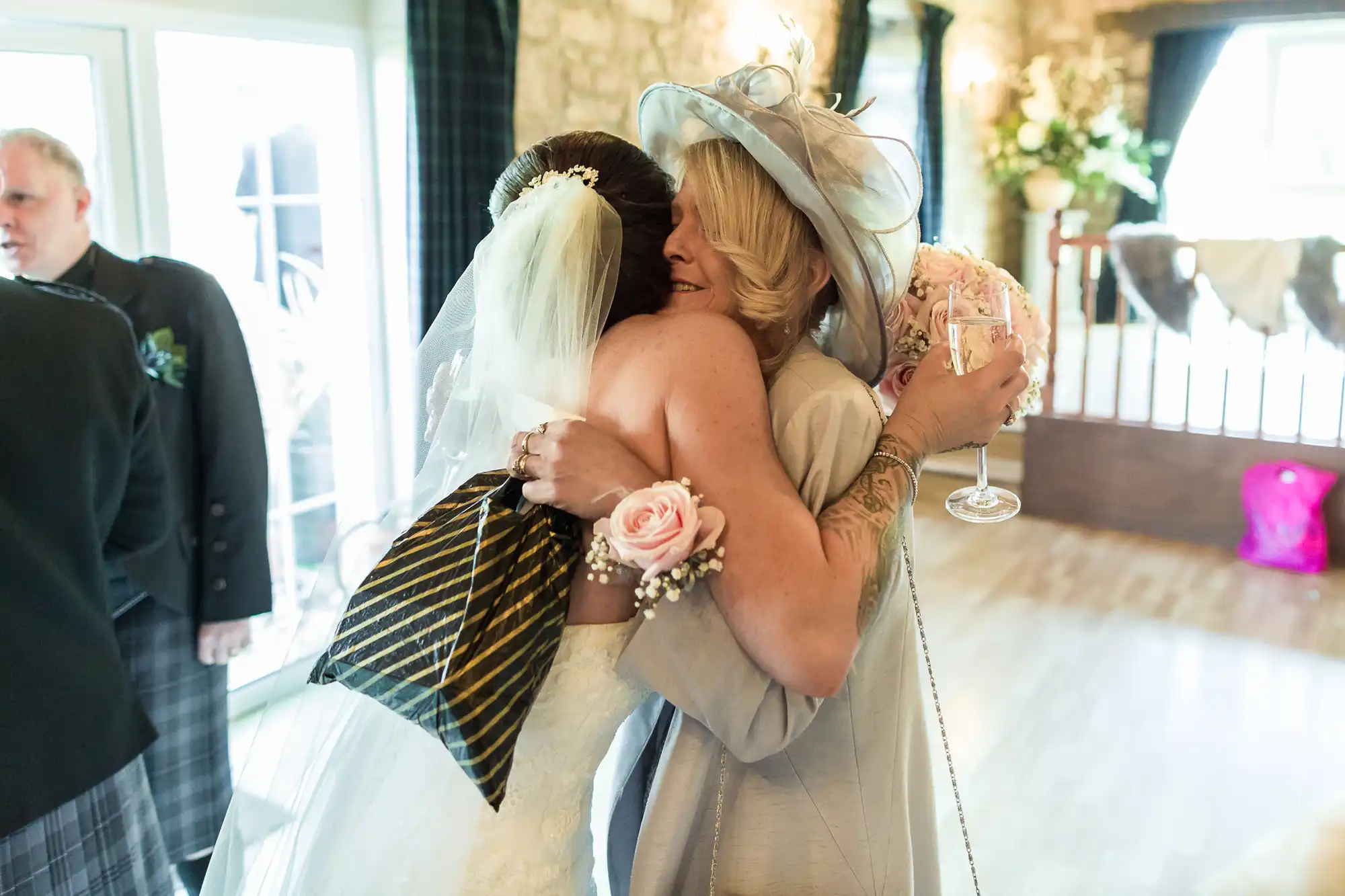 A bride in a white dress and veil hugs a woman in a pale hat and suit, holding a glass of wine and a pink rose corsage. They embrace warmly in a bright room with wooden floors, large windows, and stone walls. Other guests are partially visible.