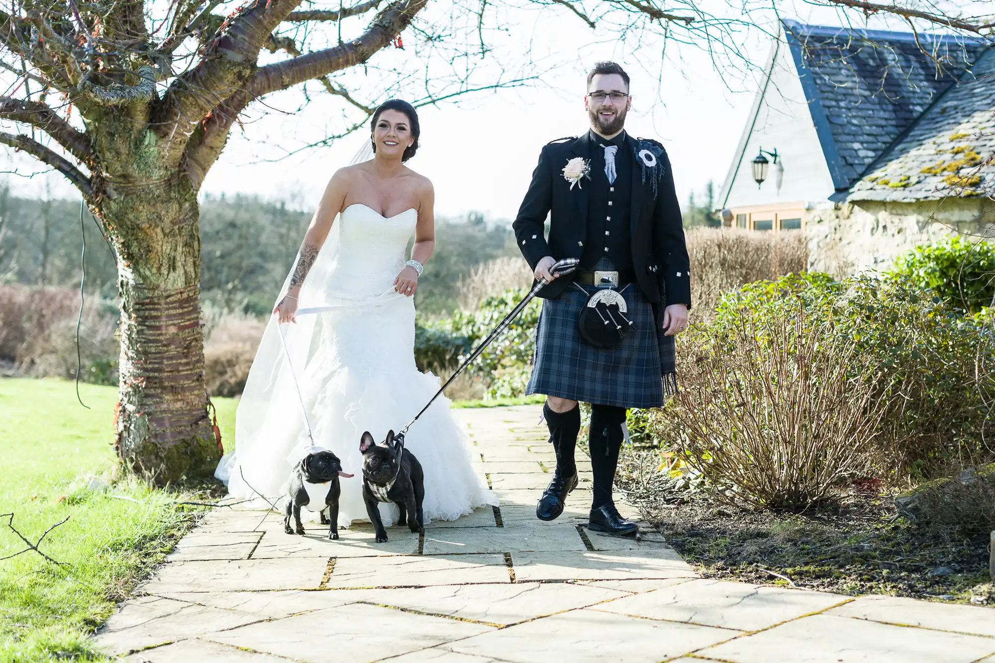 A bride in a white dress and a groom in a kilt walk two small black dogs on a stone path outdoors. The sun is shining, and they are smiling. Leafless trees and bushes surround them, with a house visible in the background.
