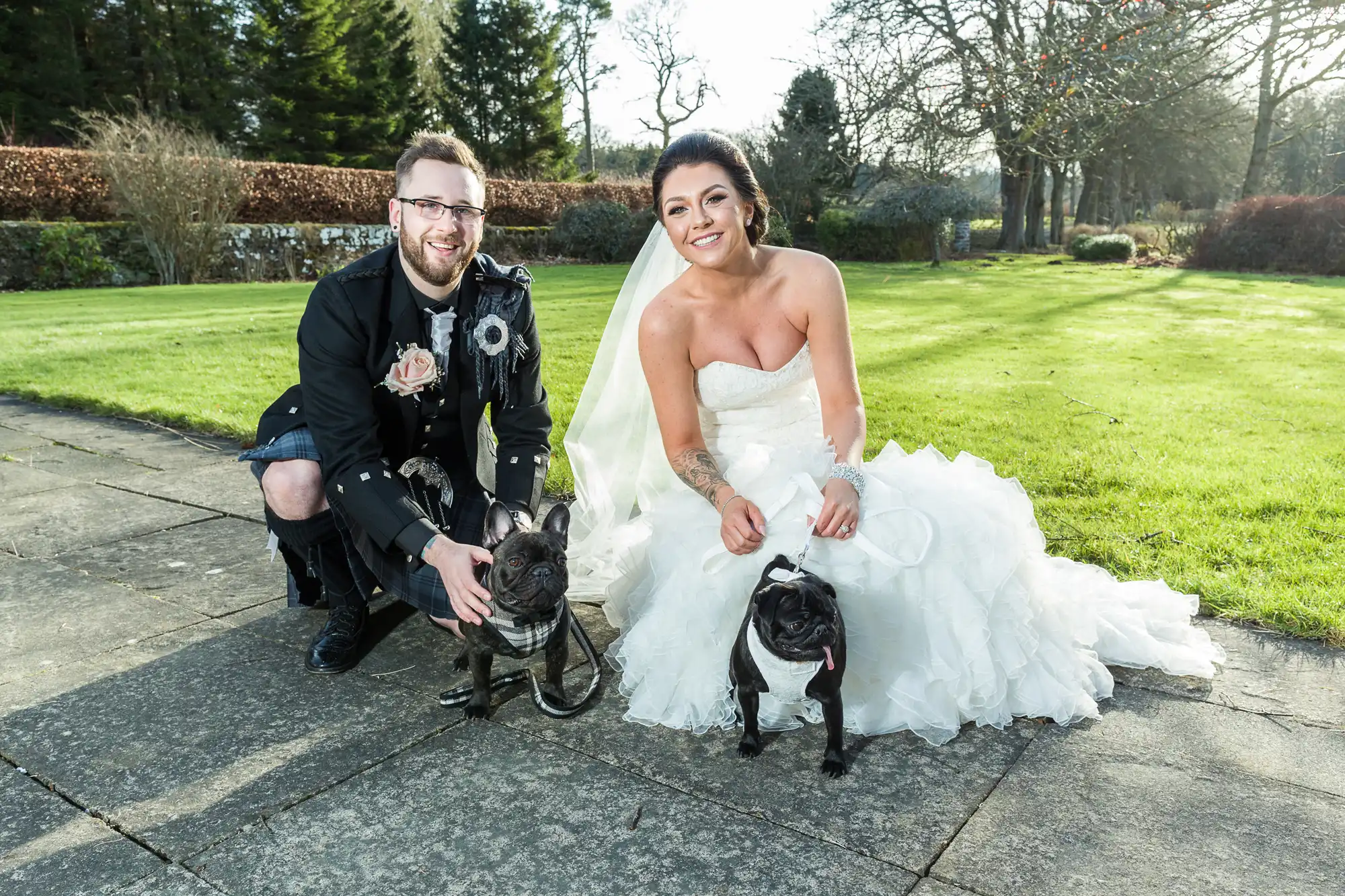 A bride in a strapless white gown and a groom in a black kilt outfit kneel on a stone path, smiling outdoors. Each holds a French bulldog on a lead. Sunlight brightens the green lawn, trees, and bushes behind them.