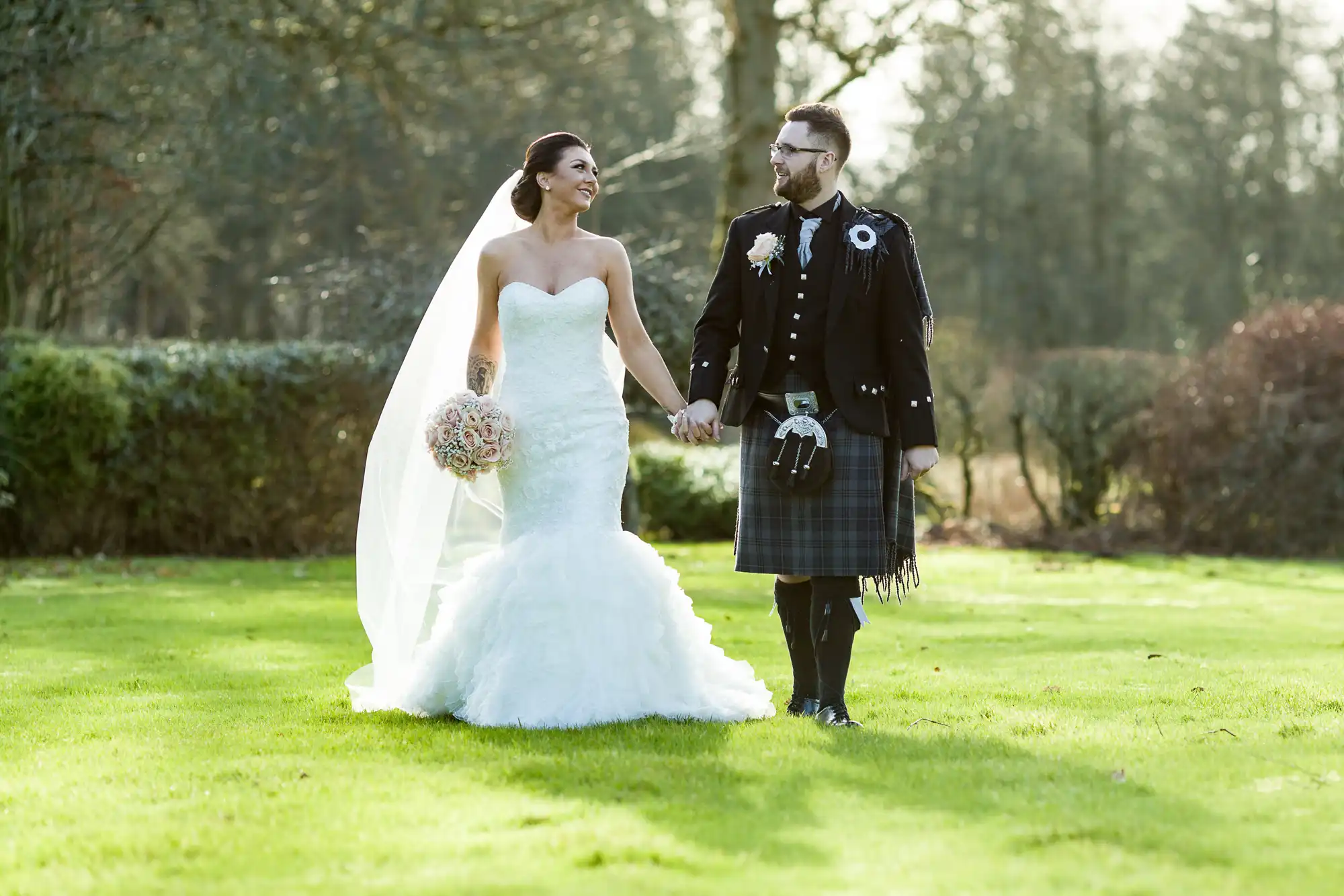 A smiling bride in a strapless white gown and veil holds a bouquet whilst walking hand in hand with a groom in a black jacket, kilt, and sporran. They look at each other, standing on green grass with trees and shrubs in the background.