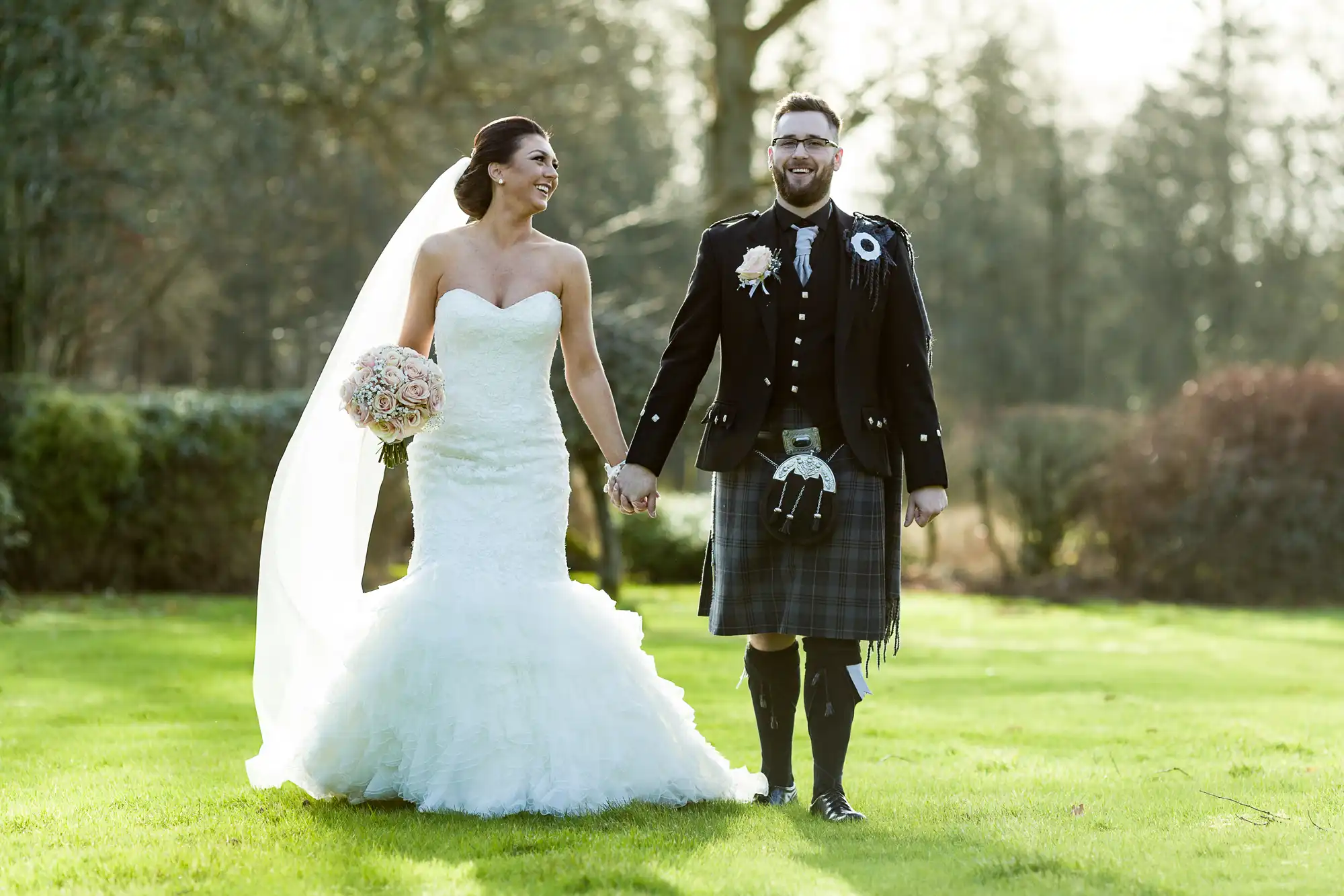 A bride in a strapless white gown and veil holds a bouquet and smiles, holding hands with a groom in a traditional Scottish kilt and jacket, standing together on a bright green lawn with trees in the background.