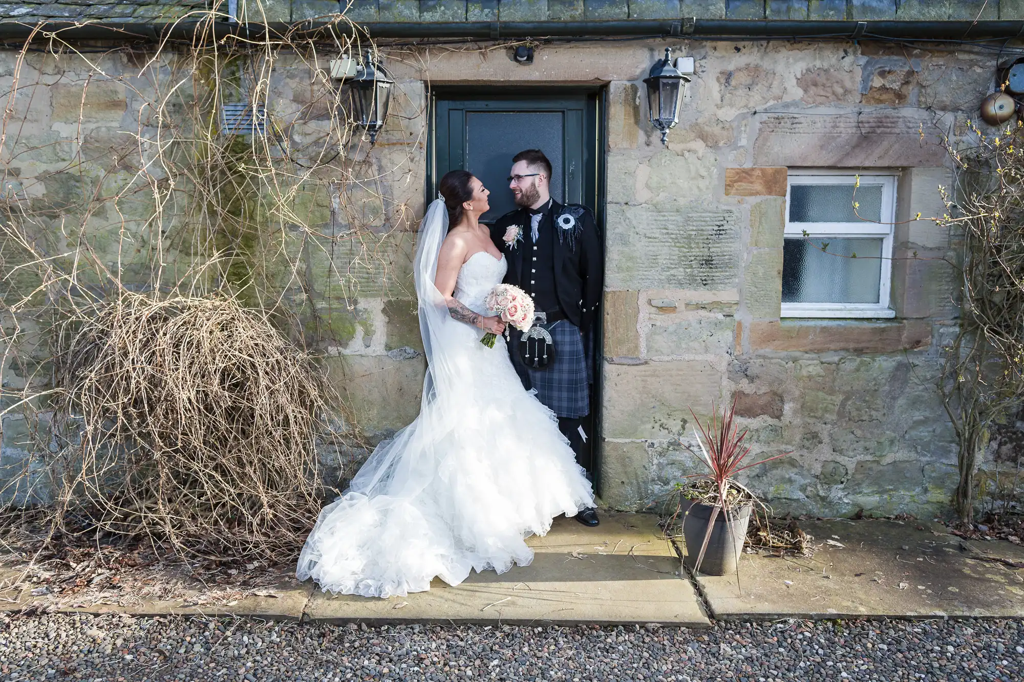 A bride in a white gown and veil holds a bouquet and smiles at a groom in a black kilt outfit. They stand close together in front of a rustic stone building with a dark door, lanterns, leafless vines, and a potted plant.
