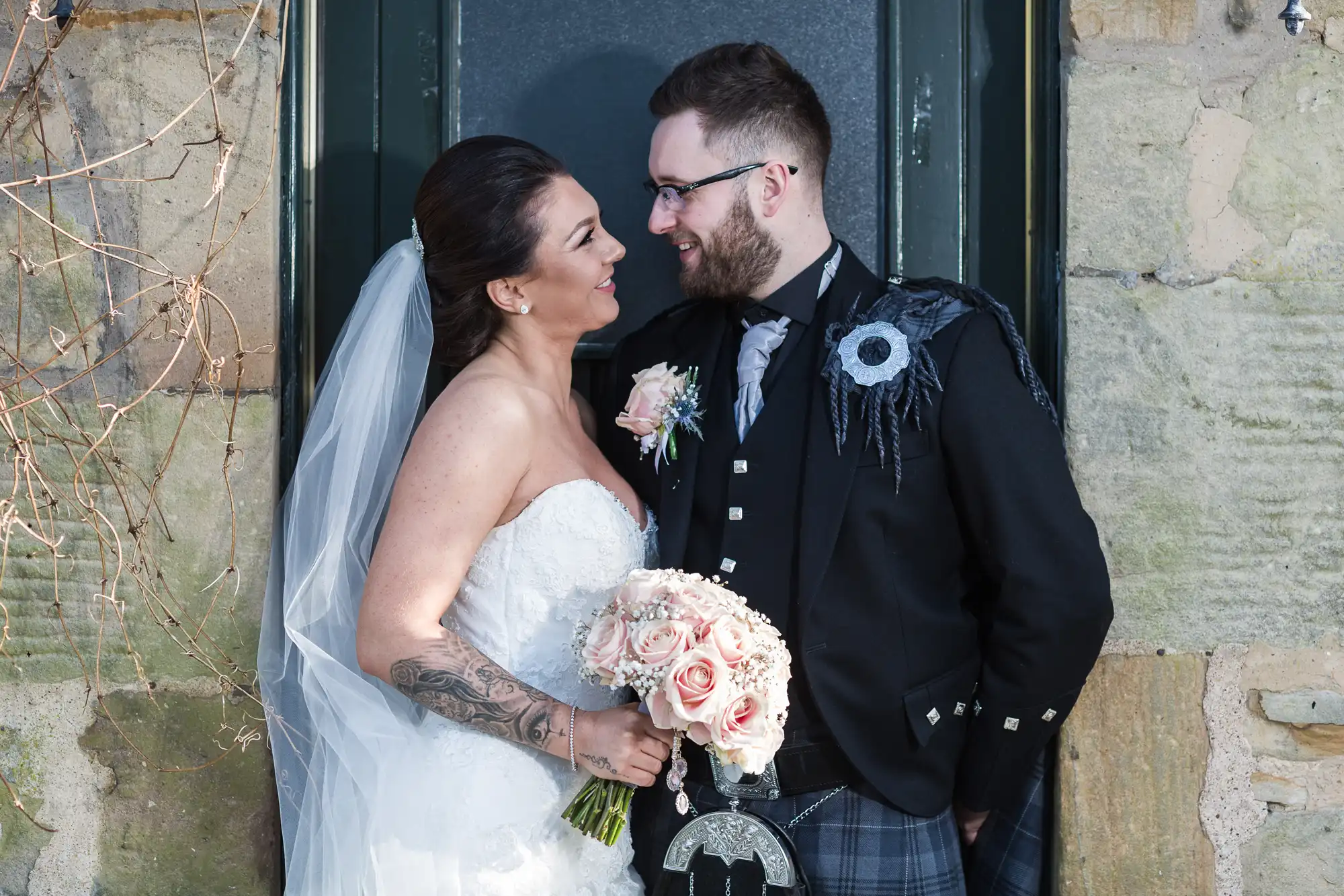 A bride in a strapless white gown and veil, holding a pink rose bouquet, smiles at a groom in a black jacket, kilt, and sporran. They stand close together, looking at each other lovingly, in front of a stone wall and dark door.