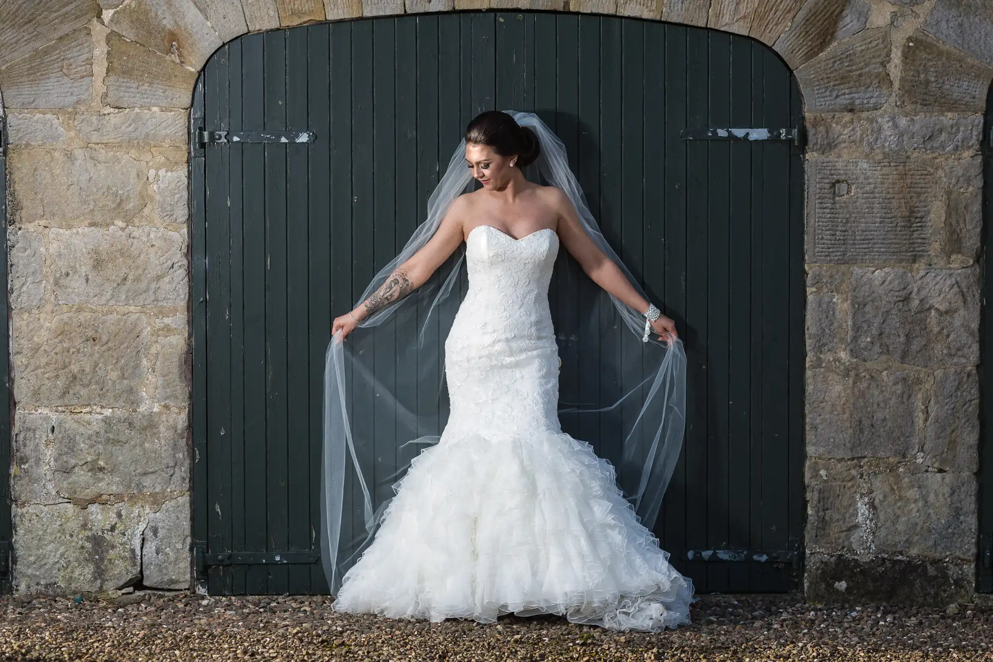 A bride in a strapless, fitted white gown with a flared ruffled skirt stands in front of dark green wooden doors. She holds out her long veil and smiles, with her hair up and a tattoo visible on her forearm. Stone walls frame the doorway.