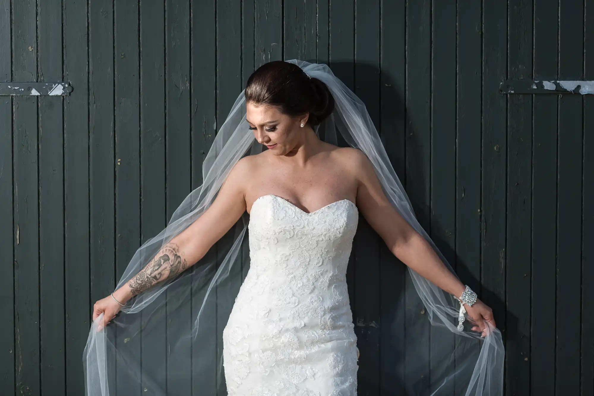 The Barn at Harburn wedding photographer image of a bride in a strapless, lace wedding dress stands against a dark wooden wall, holding her sheer veil out to each side. She has dark hair in an updo, a tattoo on her left arm, and is looking down with a gentle expression.