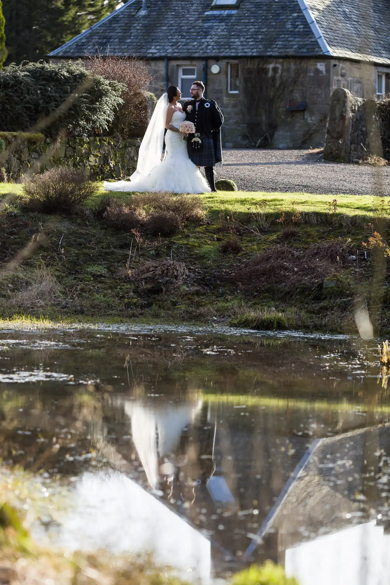 A bride in a white dress and a groom in traditional Scottish attire stand together outdoors, holding hands and facing each other, with an old stone house behind them and their reflection visible in a pond in the foreground.