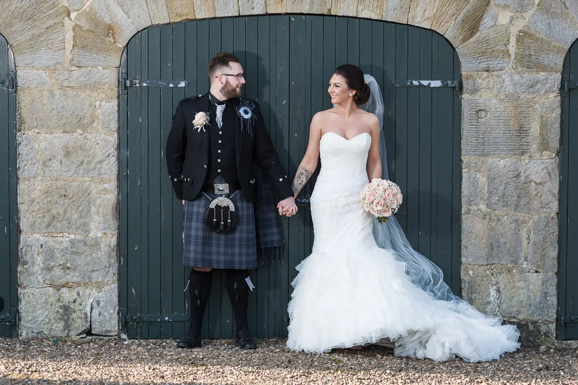 A groom in traditional Scottish attire and a bride in a strapless white gown hold hands and smile at each other, standing in front of a large arched wooden door set in a stone wall. The bride holds a bouquet of pale pink flowers.