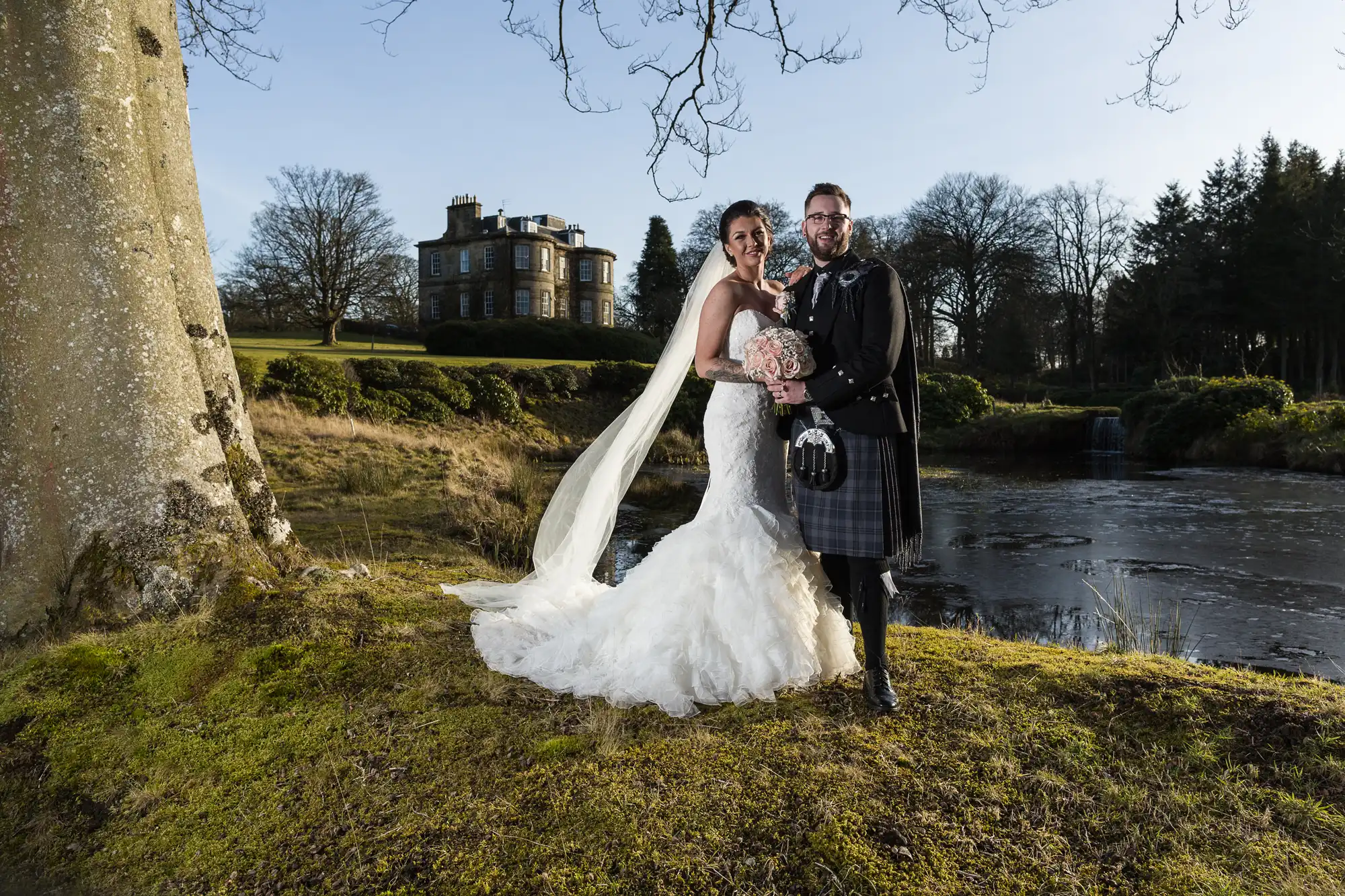 A bride in a white gown and veil stands next to a groom in a traditional Scottish kilt by a tree, smiling and holding a bouquet. Behind them is a large manor house, frozen pond, and trees under a clear sky, suggesting a winter wedding.