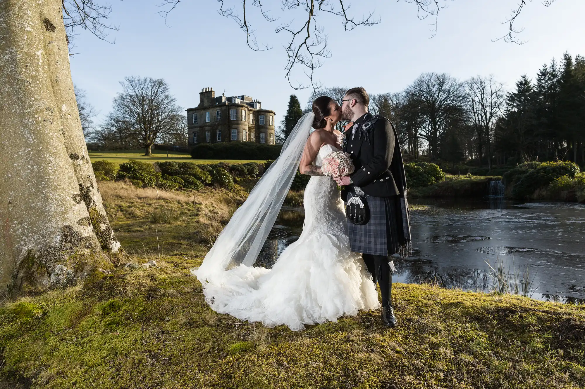 A bride in a white dress and veil and a groom in a black jacket and tartan kilt embrace by a pond, with a bouquet in hand. Behind them, there is a stately home, trees, and an expansive lawn under a clear sky.
