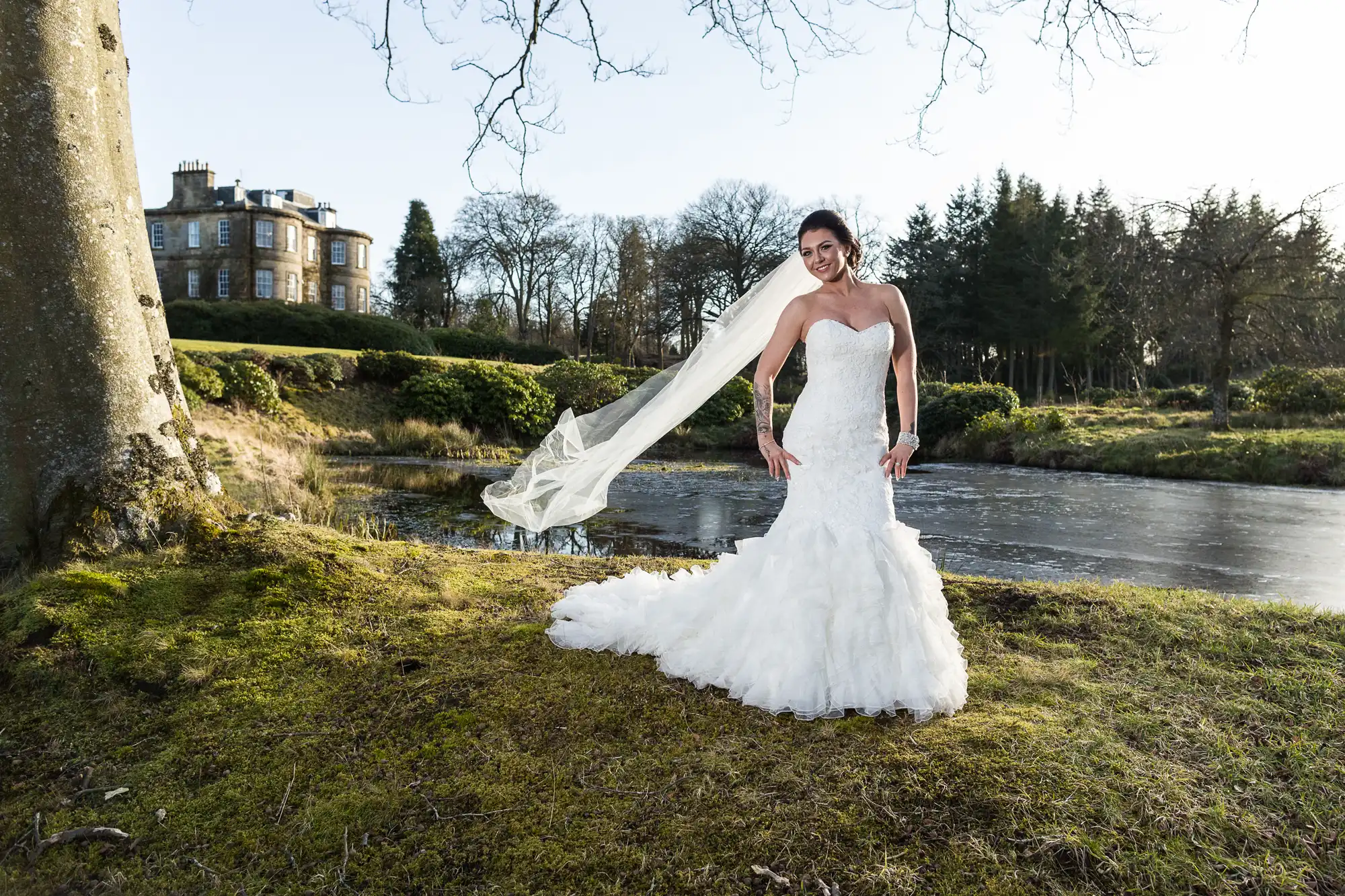 A smiling bride in a strapless white gown and long veil stands outdoors on grass near a pond, with trees and a large stone building in the background. The scene is bright and serene, suggesting a chilly but sunny day.