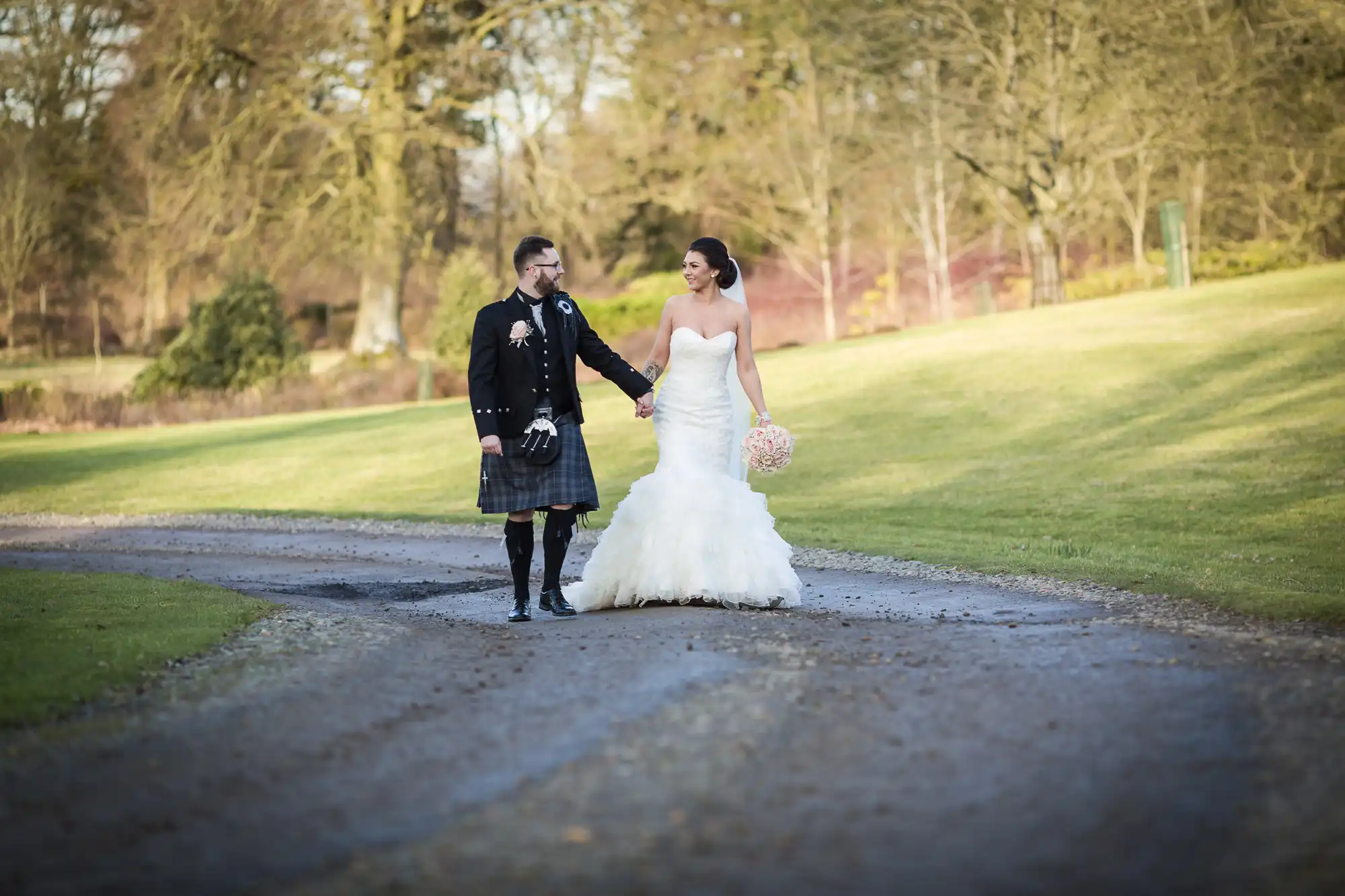 A bride in a strapless white gown holds a bouquet and walks hand-in-hand with a groom wearing a black jacket, traditional Scottish kilt, and sporran. They smile at each other on a winding path in a sunlit park with green grass and trees.