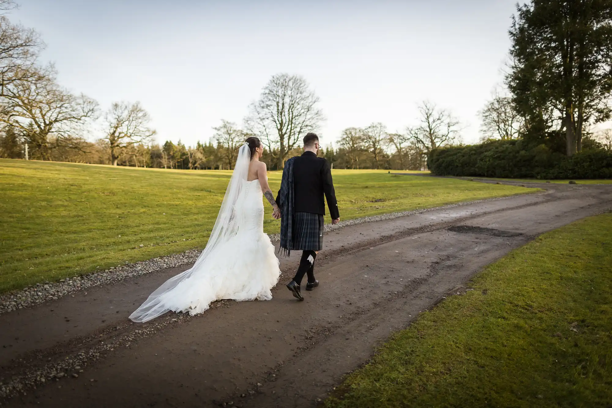A bride in a white wedding dress with a long veil walks hand in hand with a groom wearing a dark jacket and tartan kilt along a curved path in a grassy, tree-lined park on a clear day. Both are walking away from the camera.