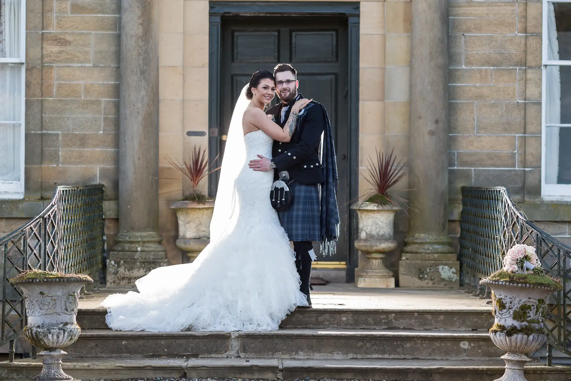 A bride in a white strapless gown and a groom in a traditional Scottish kilt embrace and smile on the stone steps of a grand building with tall columns and large windows, flanked by decorative planters and ornate railings.