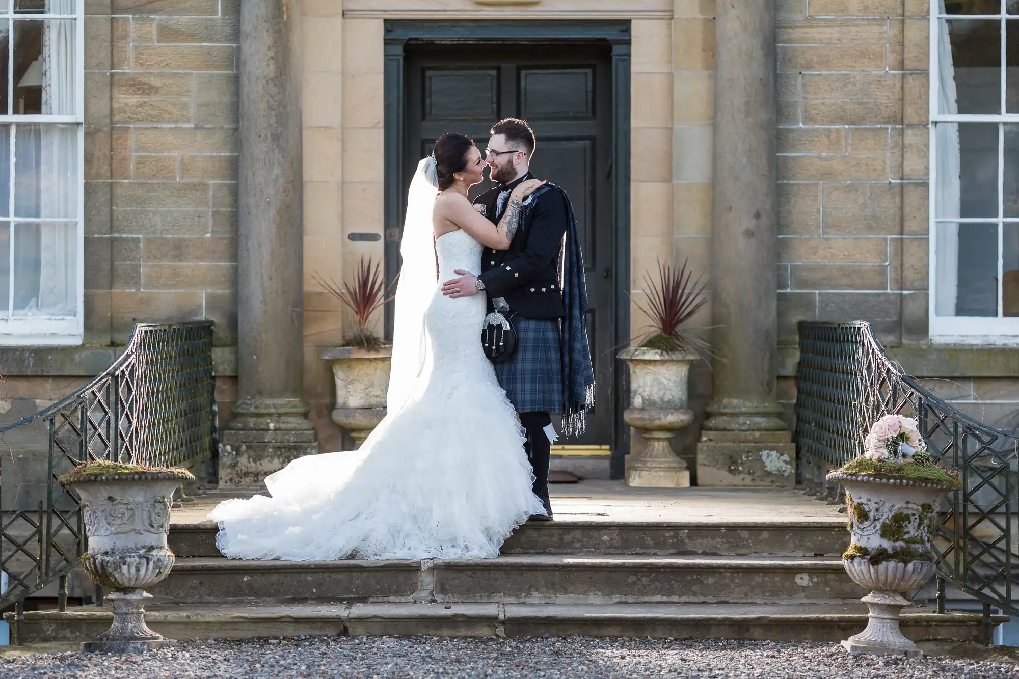 A bride in a white gown and veil embraces a groom in traditional Scottish attire, including a kilt, on stone steps outside an historic building with pillars, large door, and potted plants on either side. Both smile lovingly at each other.