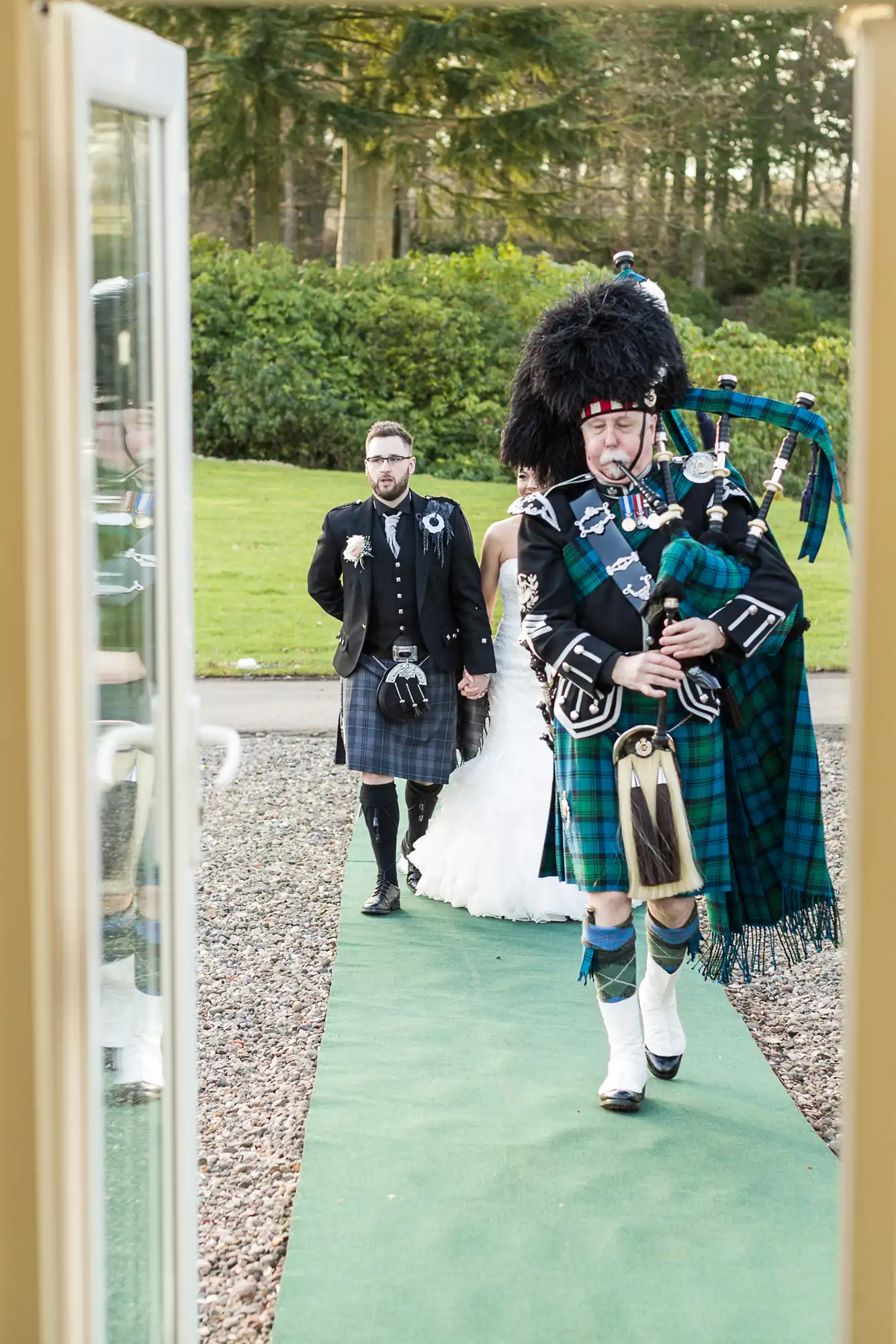A bagpiper in traditional Scottish attire leads a bride and groom along a green outdoor aisle. The groom and bagpiper wear kilts and jackets; the bride wears a white gown. Trees and grass are in the background. The scene is viewed from an open doorway.