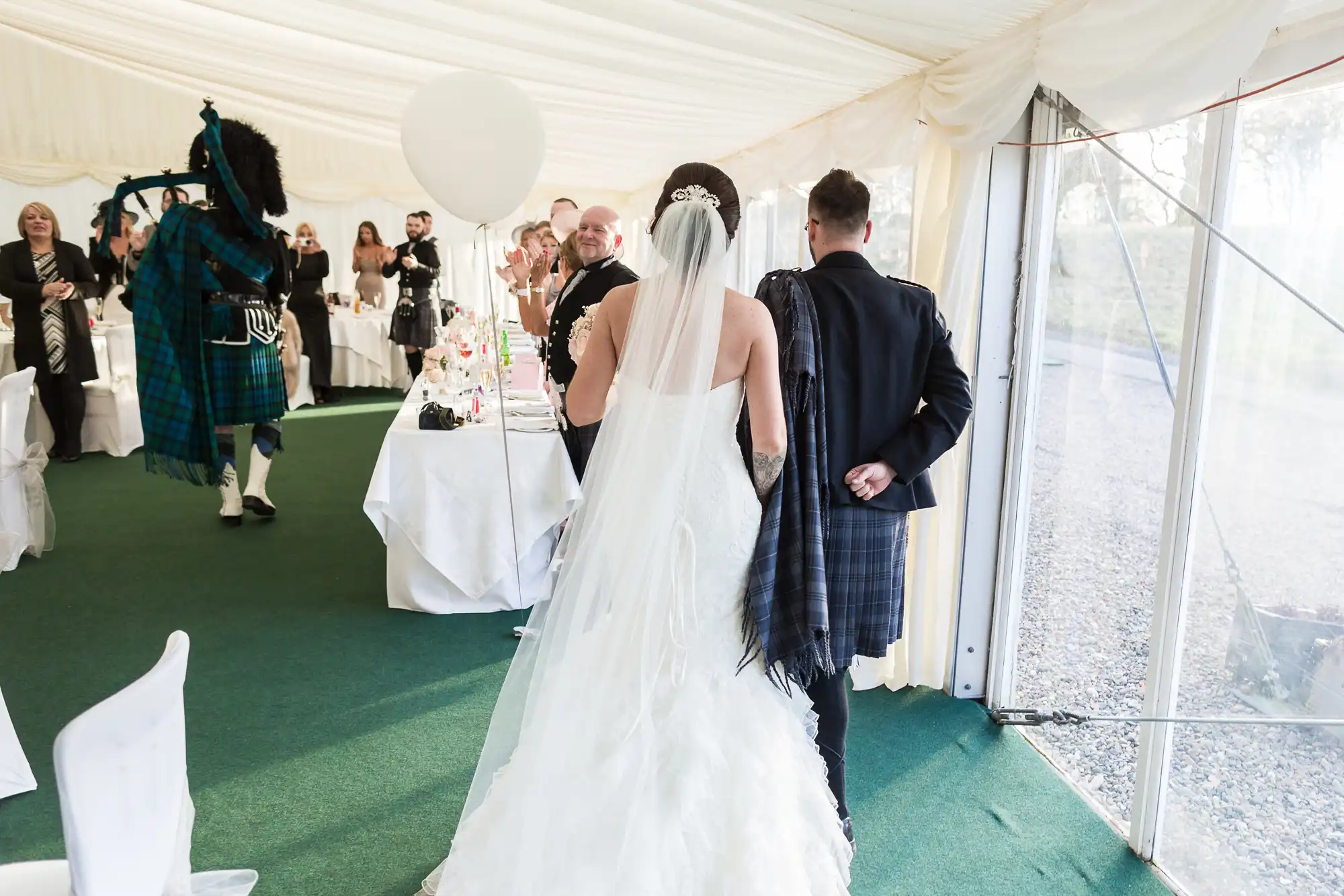 A bride in a white dress and veil walks with a groom in Scottish attire into a bright marquee, guests standing and applauding. A bagpiper in traditional tartan plays near decorated tables with a wedding cake visible. Sunlight streams through clear windows on the right.
