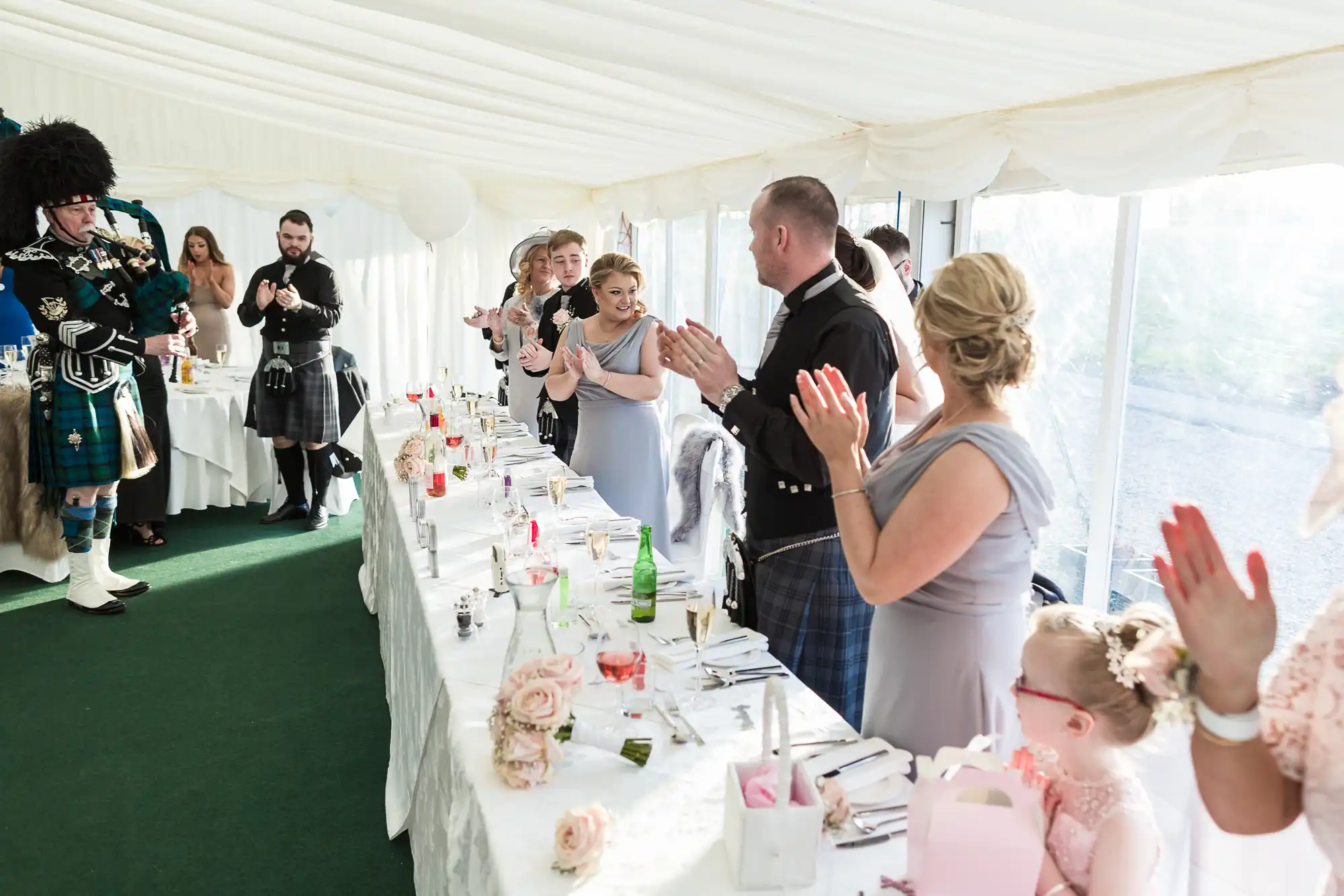 A wedding reception in a bright marquee shows guests in formal attire, including kilts and dresses, applauding around a long table decorated with flowers and drinks. A bagpiper in traditional Scottish dress stands to the left, whilst a young girl with glasses sits in the foreground.