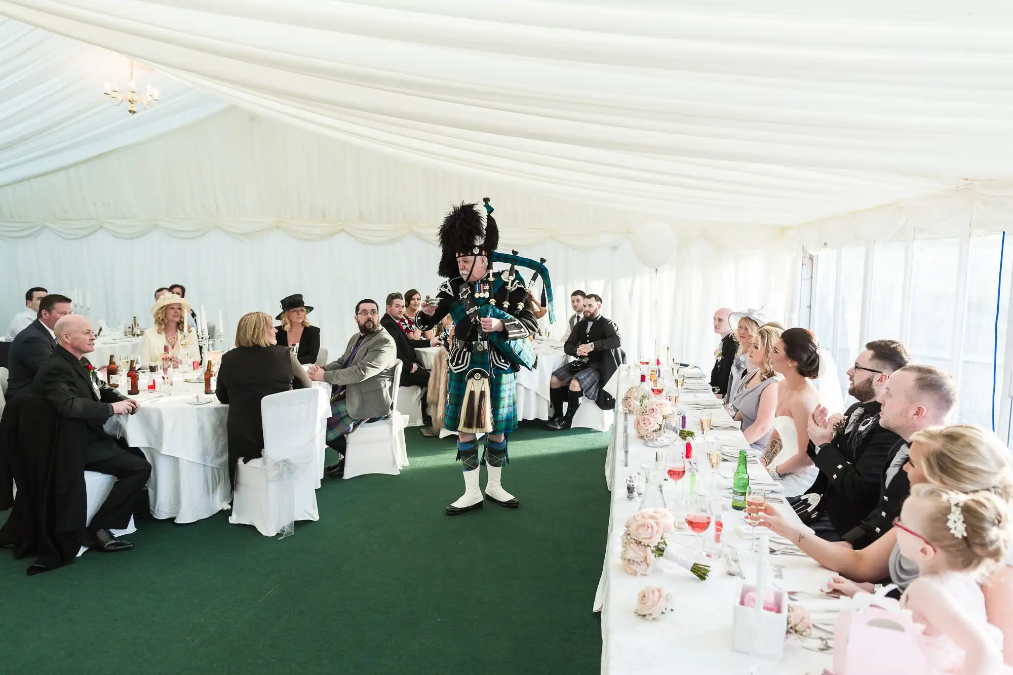A bagpiper in traditional Scottish attire walks between two long tables of wedding guests seated in a bright, white marquee. The tables are decorated with flowers and glasses. Guests watch the musician, some smiling and dressed in formal or semi-formal clothing.