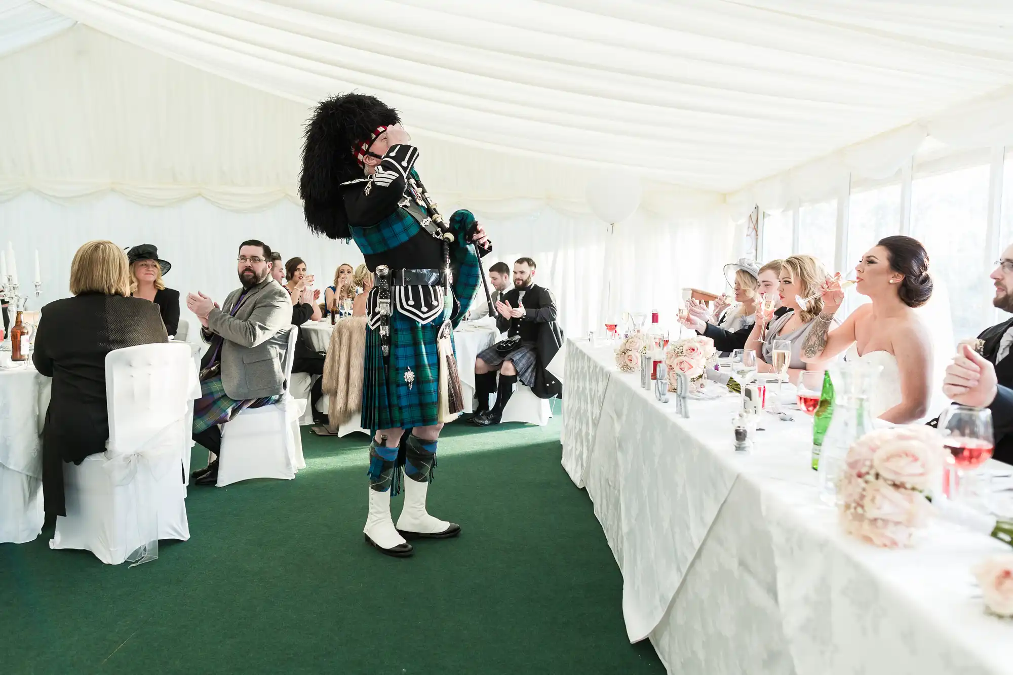 A man in traditional Scottish attire, including a kilt and feathered hat, plays bagpipes in a white marquee at a wedding reception, while seated guests clap and smile at long, decorated tables with white cloths and floral centrepieces.