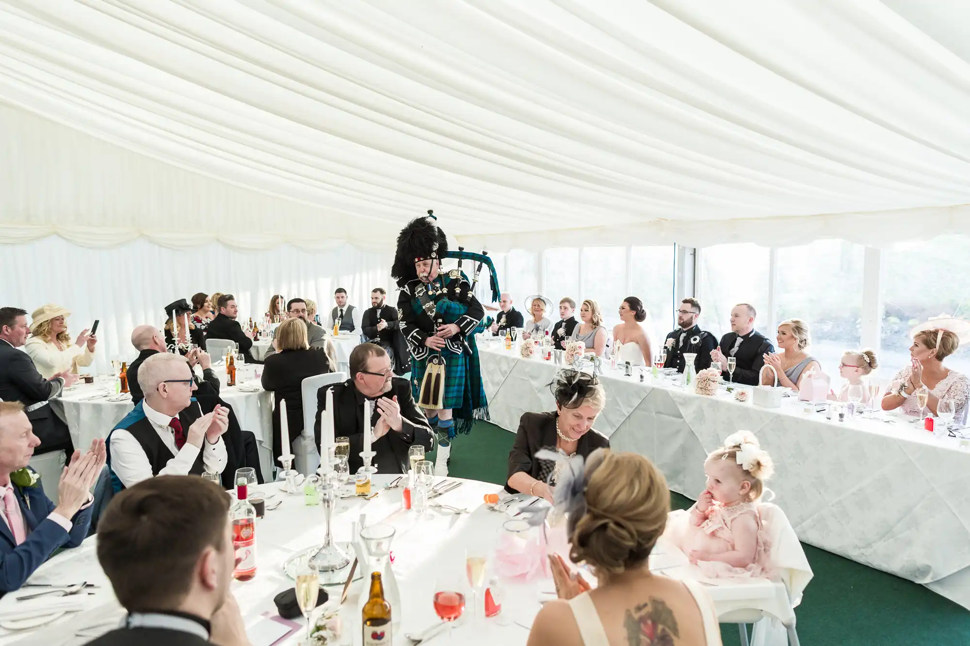 A wedding reception in a white marquee shows guests seated at long tables, applauding as a bagpiper in traditional Scottish attire enters. People of all ages, dressed formally, are engaged and smiling, with daylight streaming through the marquee’s windows.