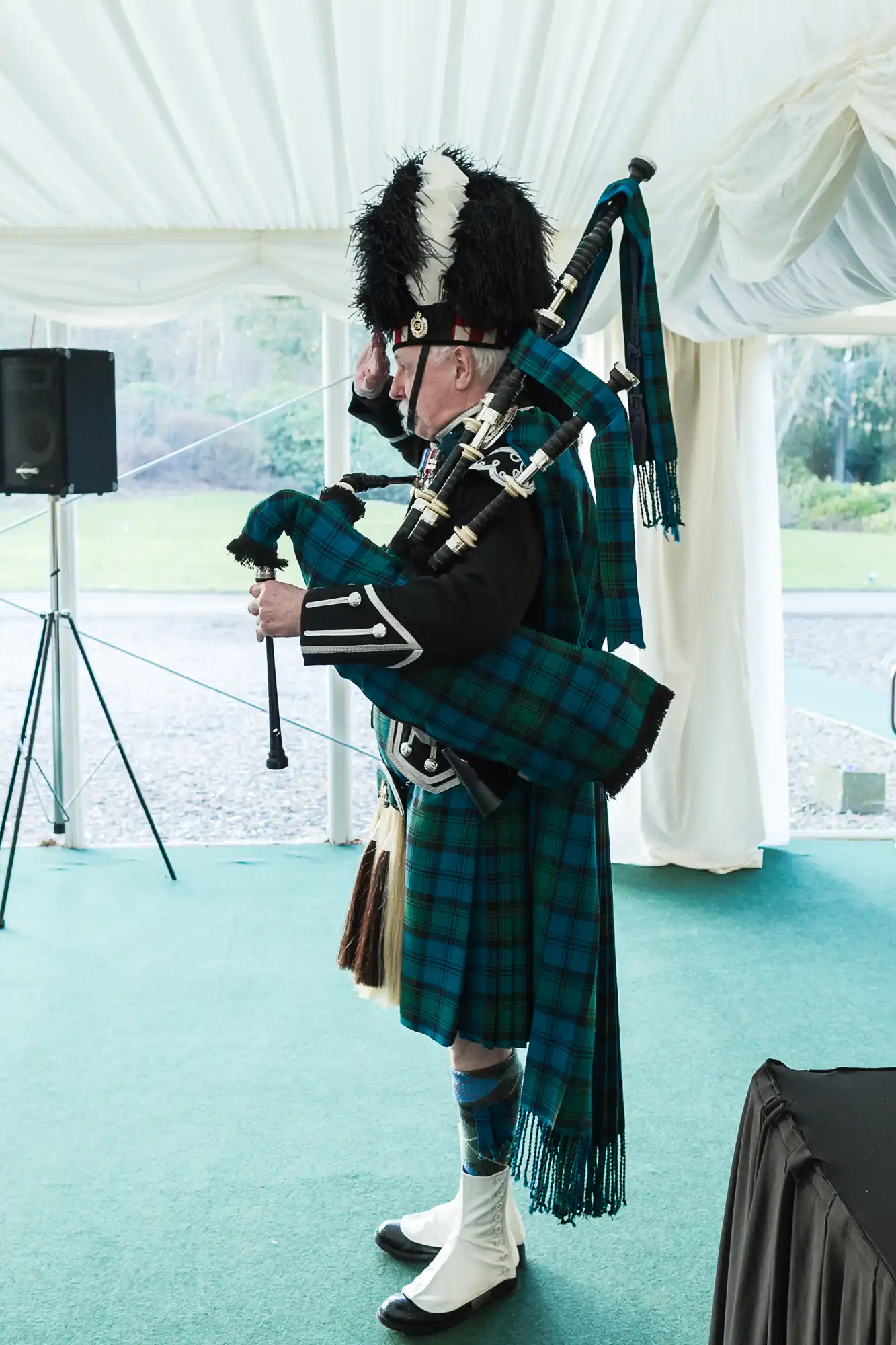 A man in traditional Scottish attire plays bagpipes indoors. He wears a feathered hat, black jacket, blue-green tartan kilt with matching sash, white spats, and blue socks. He stands on green carpet near a speaker and large window, saluting with his right hand.
