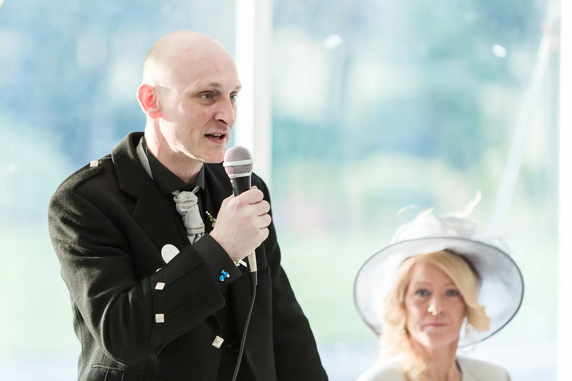 A bald man in a black suit speaks into a microphone, wearing a patterned tie and a buttonhole. Next to him, a blonde woman in a white hat and light-coloured outfit sits, slightly out of focus. Bright, natural light streams in from large windows behind them.
