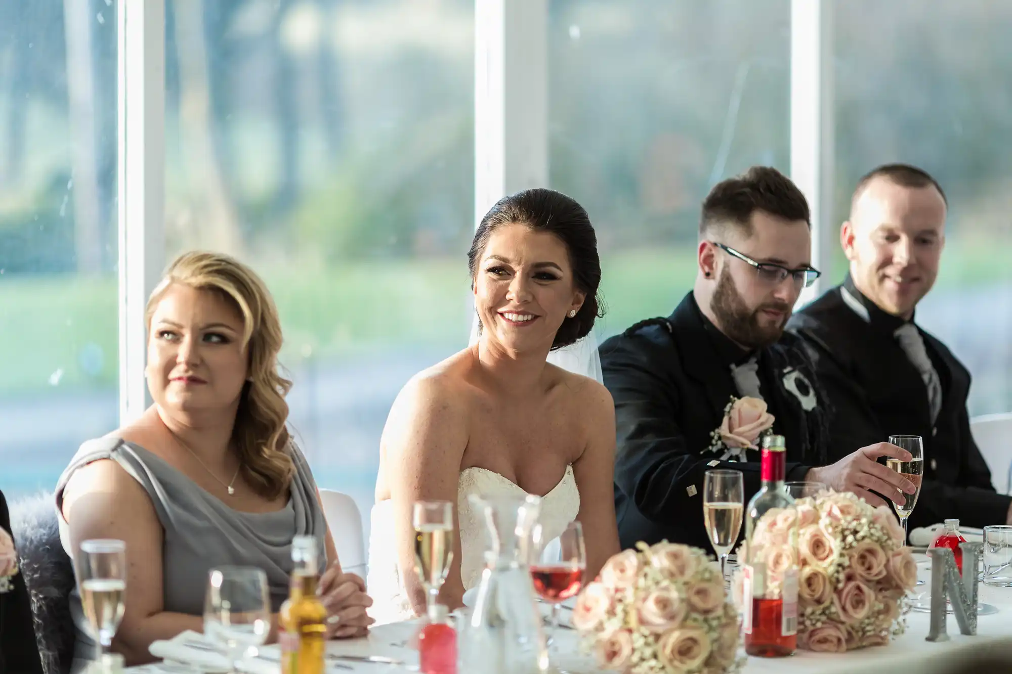 Four people sit at a wedding reception table decorated with blush pink flower bouquets and drinks. The bride, in a white dress, smiles in the centre. Two men in suits and a woman in a grey dress sit beside her. Sunlight streams through large windows behind them.