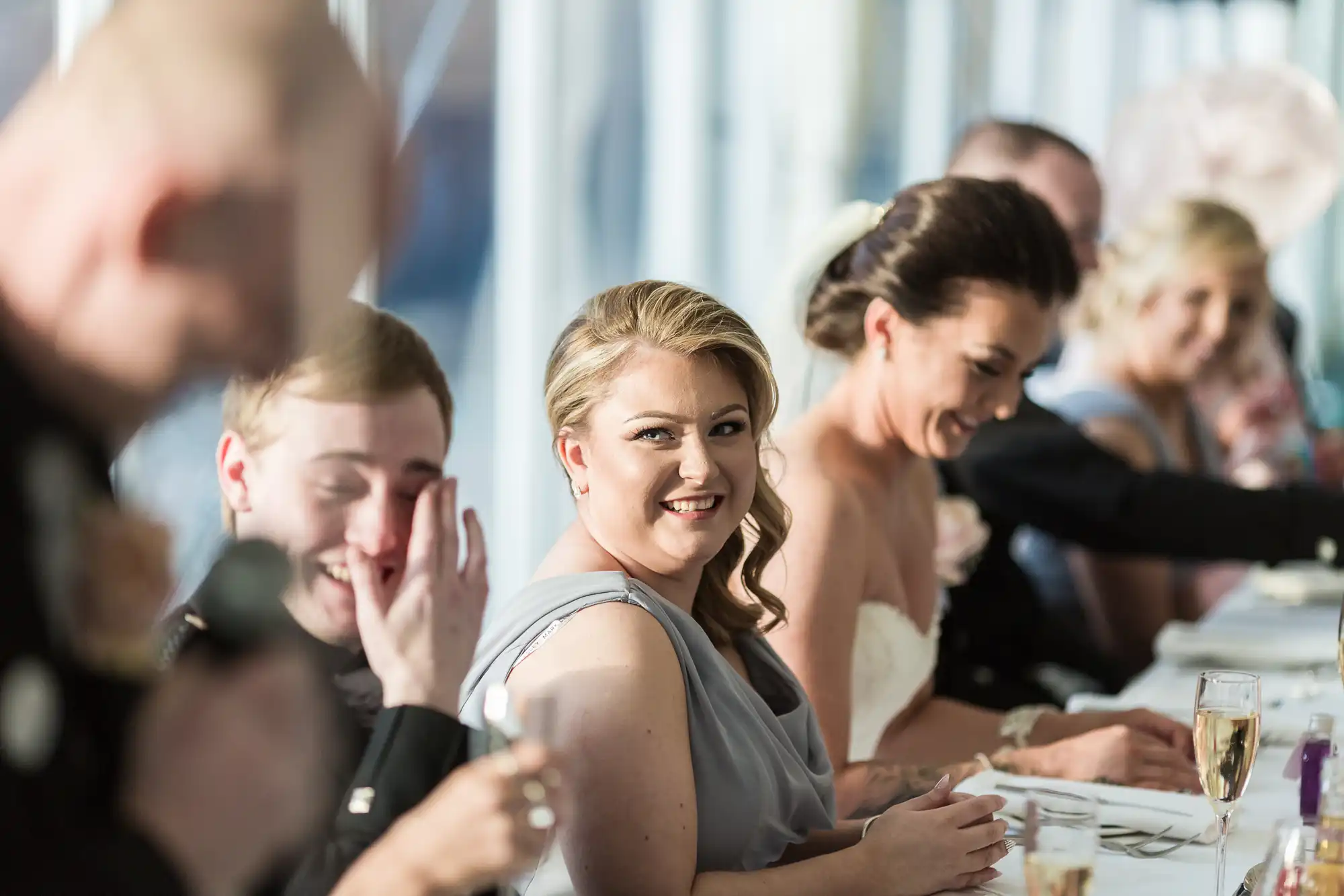 A woman in a grey dress smiles at the camera while seated at a formal table with others. The group appears to be enjoying a joyful moment, with some people laughing. Sunlight streams through large windows in the bright background.