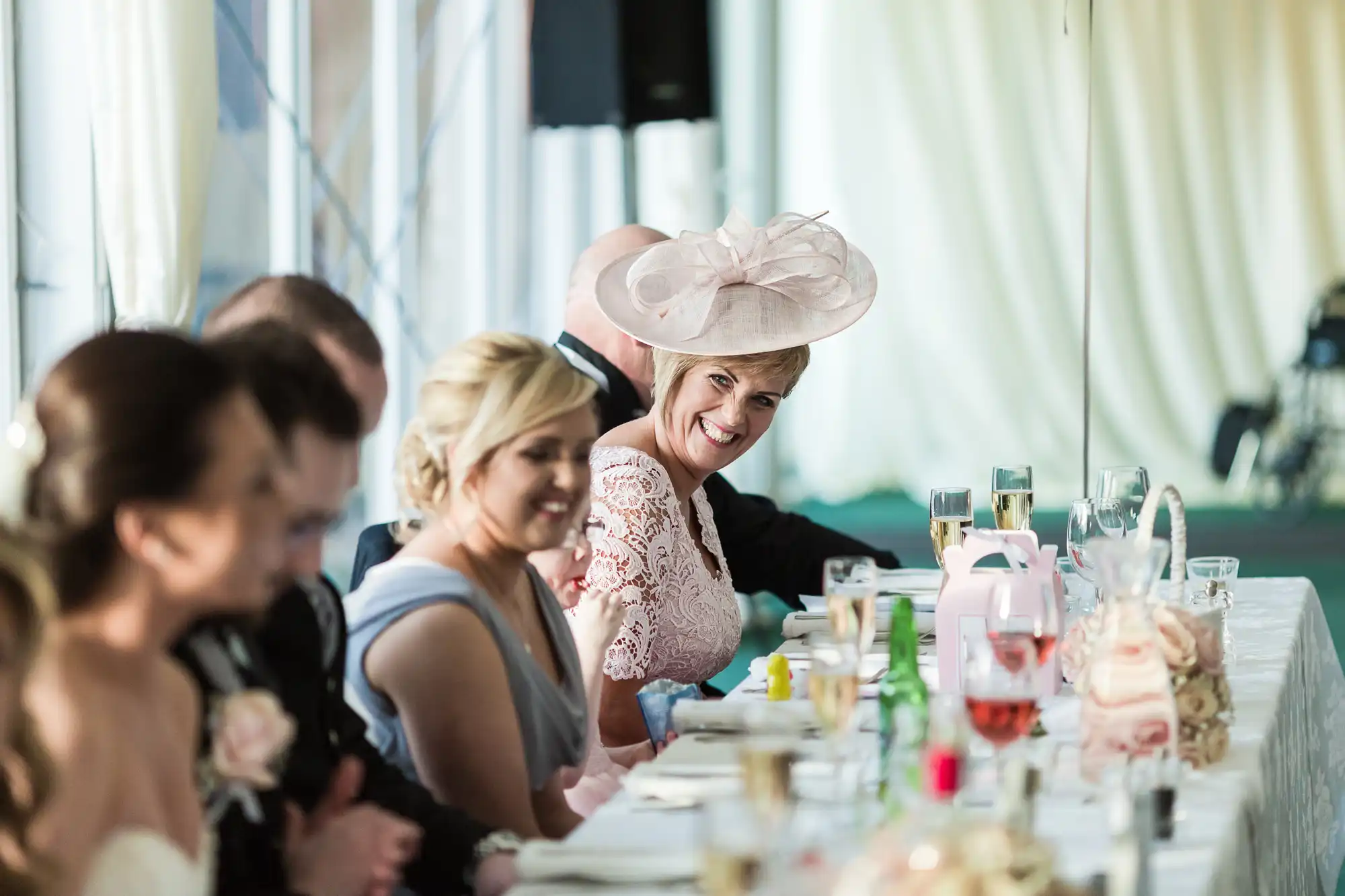 A group of people dressed formally sit at a long table during a wedding reception. One woman in a pink lace dress and a decorative hat smiles warmly. Glasses, bottles, and plates decorate the table, and soft natural light fills the room.