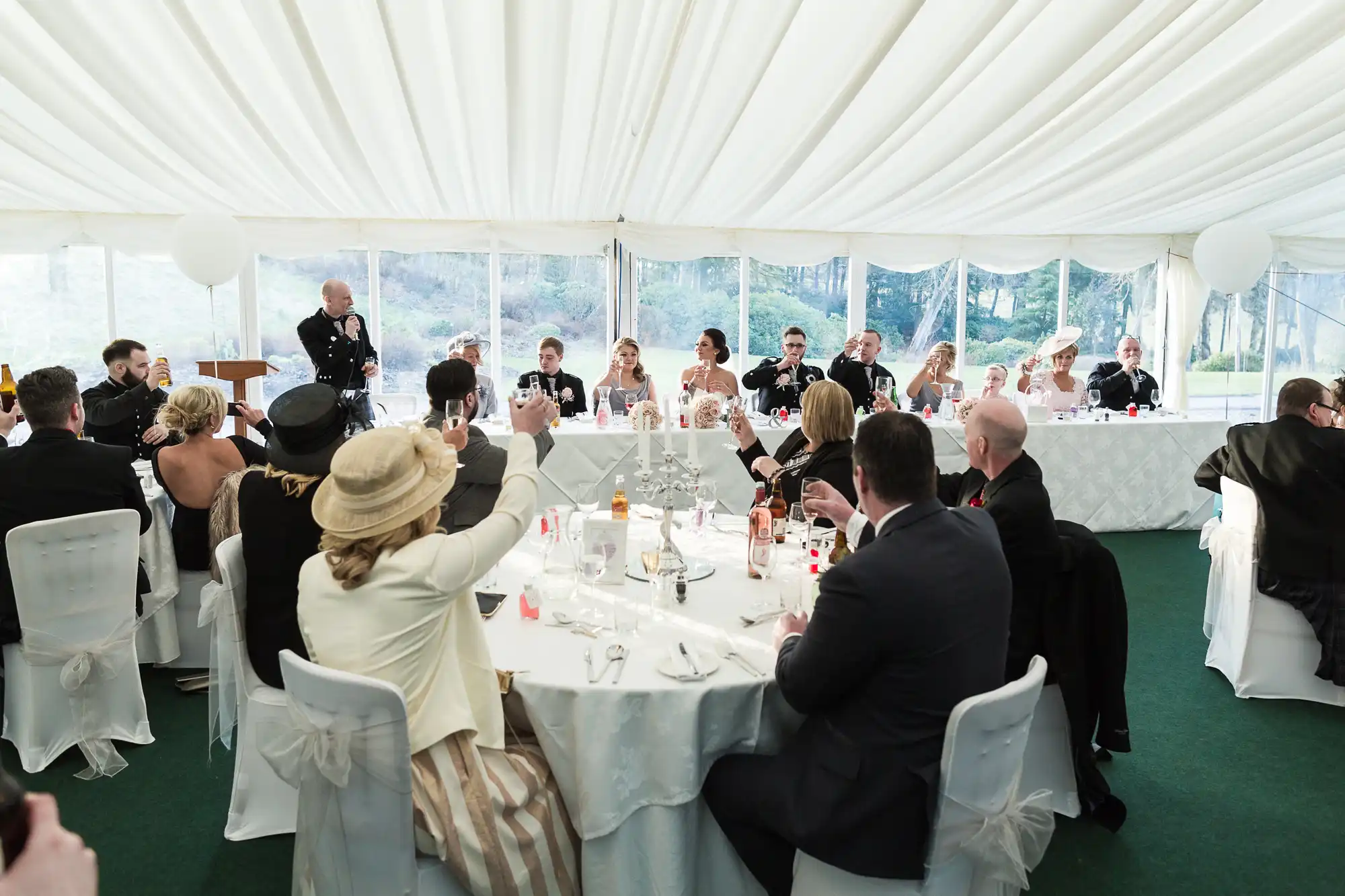 Guests seated at round tables in a white marquee raise glasses for a toast towards a long top table, where the bridal party sits. The tables are decorated simply, and daylight streams through the marquee windows, creating a festive atmosphere.