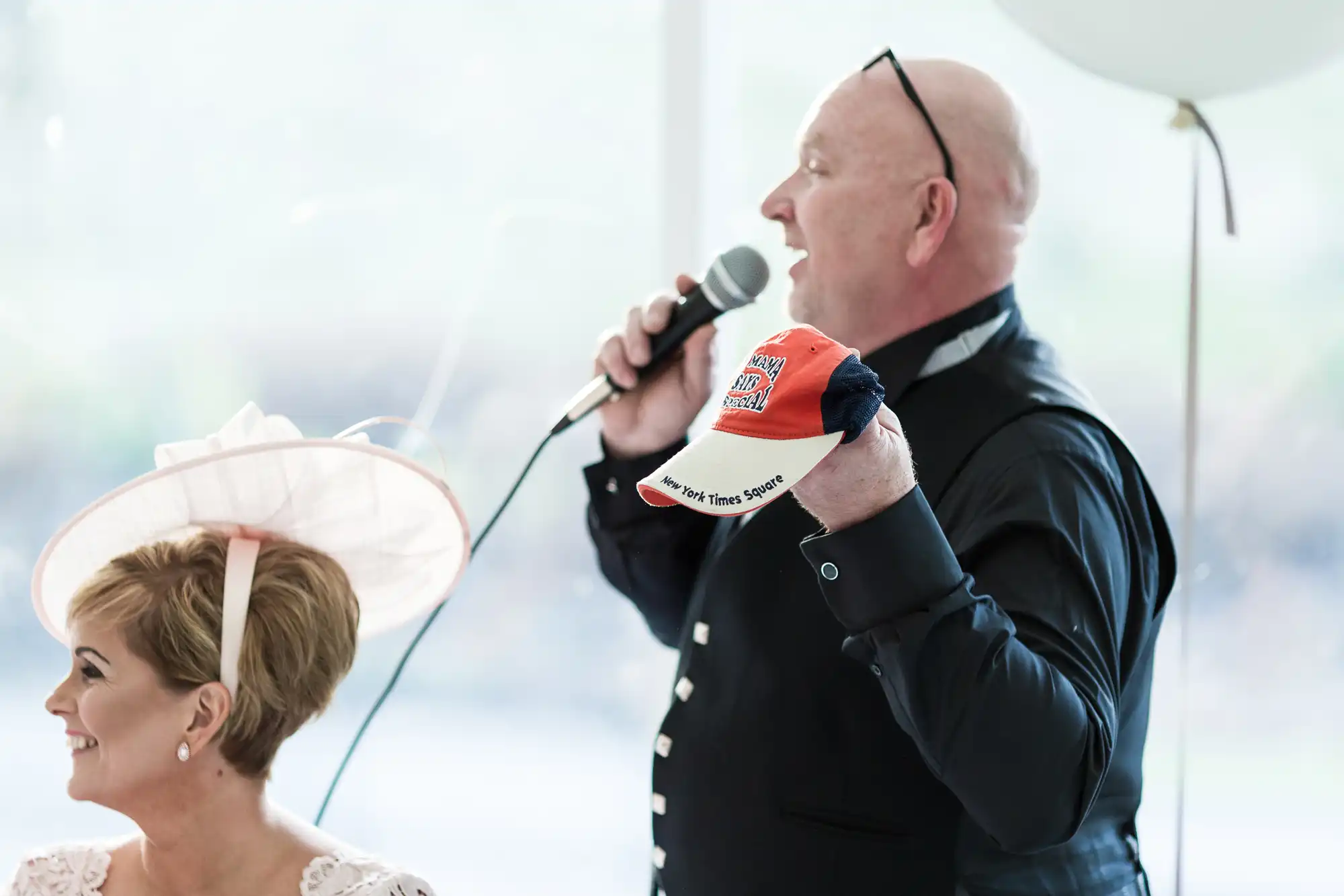 A bald man in a dark waistcoat holds a microphone and an orange cap whilst speaking at an event. Beside him, a smiling woman with short hair wears a light pink dress and an elaborate hat. The background is softly blurred with light, pastel tones.