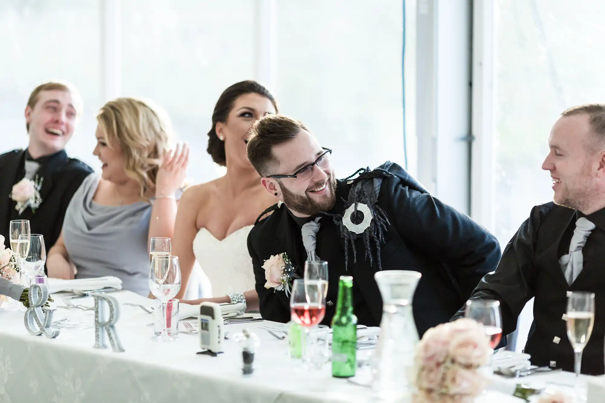 A bride in a strapless white dress sits smiling at a decorated table with four guests, all laughing and chatting. There are glasses of rosé, bottles, and floral decorations on the table, with large windows in the background letting in natural light.