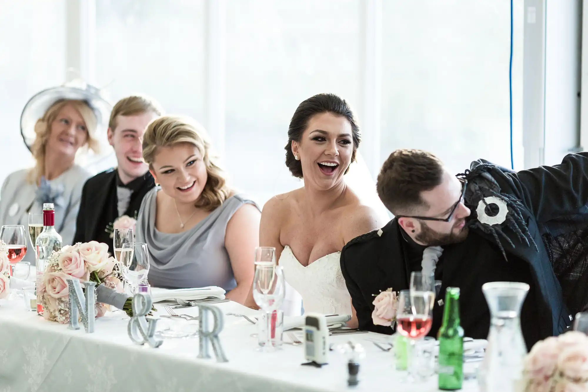 A joyful wedding party sits at a decorated table. The bride, in a strapless white dress, laughs beside a man adjusting his jacket. Two bridesmaids in grey and two men in suits also smile. Flowers, drinks, and monogrammed letters accent the bright, festive scene.