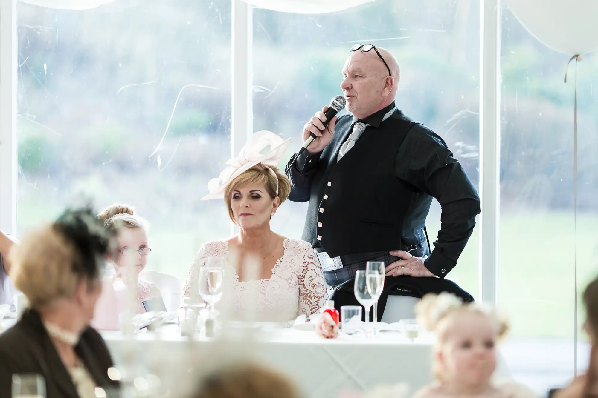 A man in formal attire stands with a microphone, speaking at a wedding reception. A woman in a light pink dress and elaborate hat sits beside him, looking forward. Guests are seated around white tables with glassware in a bright room with large windows.