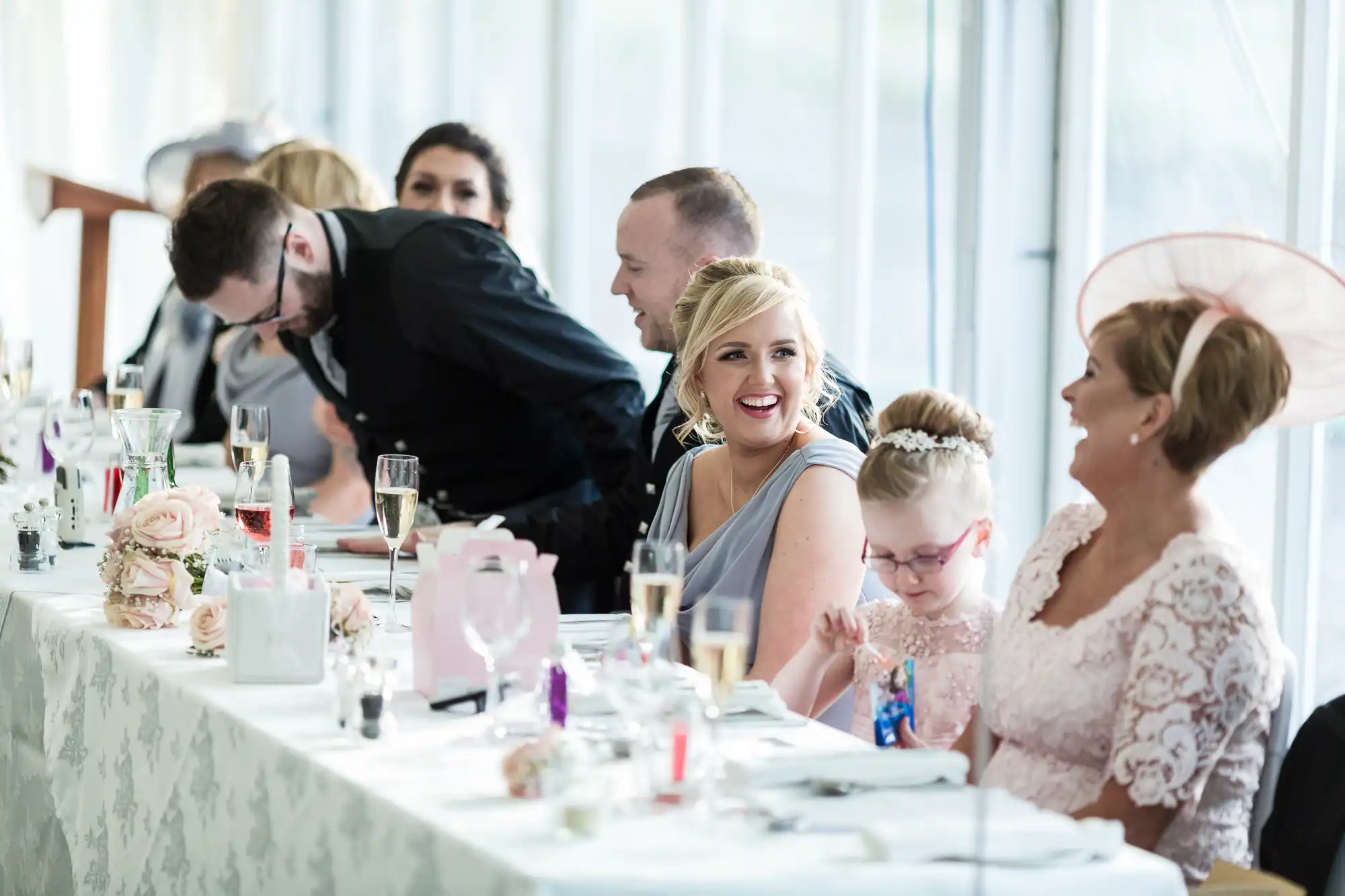 A group of people, dressed formally, sit at a long, decorated table at a wedding reception. They are talking, smiling, and laughing. The table is adorned with floral arrangements, drinks, and presents. Daylight streams in through tall windows in the background.
