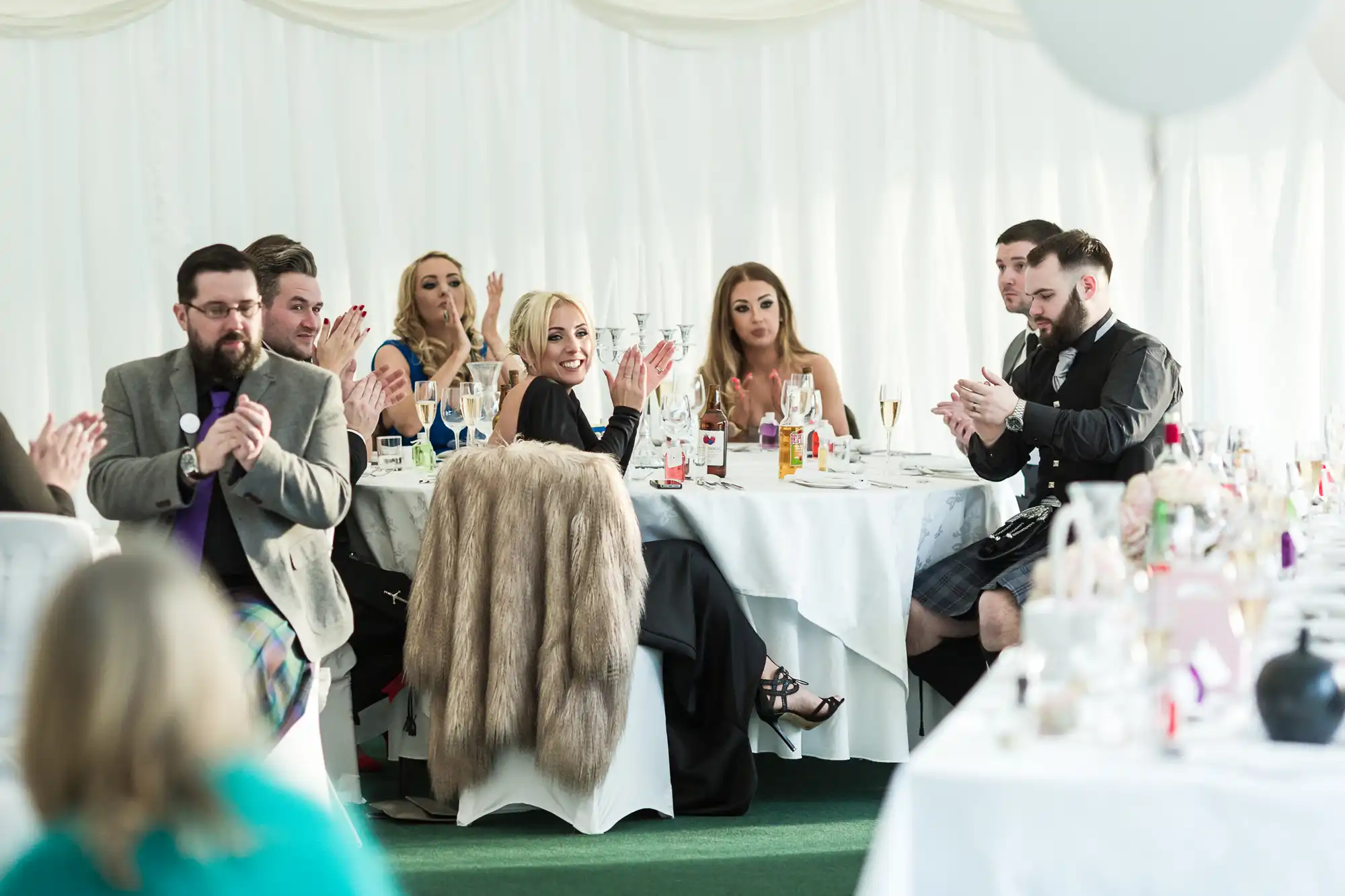 A group of people dressed formally sits at round tables, applauding and smiling. The setting appears to be a bright, indoor event with white draped walls. Elegant tableware and drinks are on the tables, and one woman wears a fur stole.