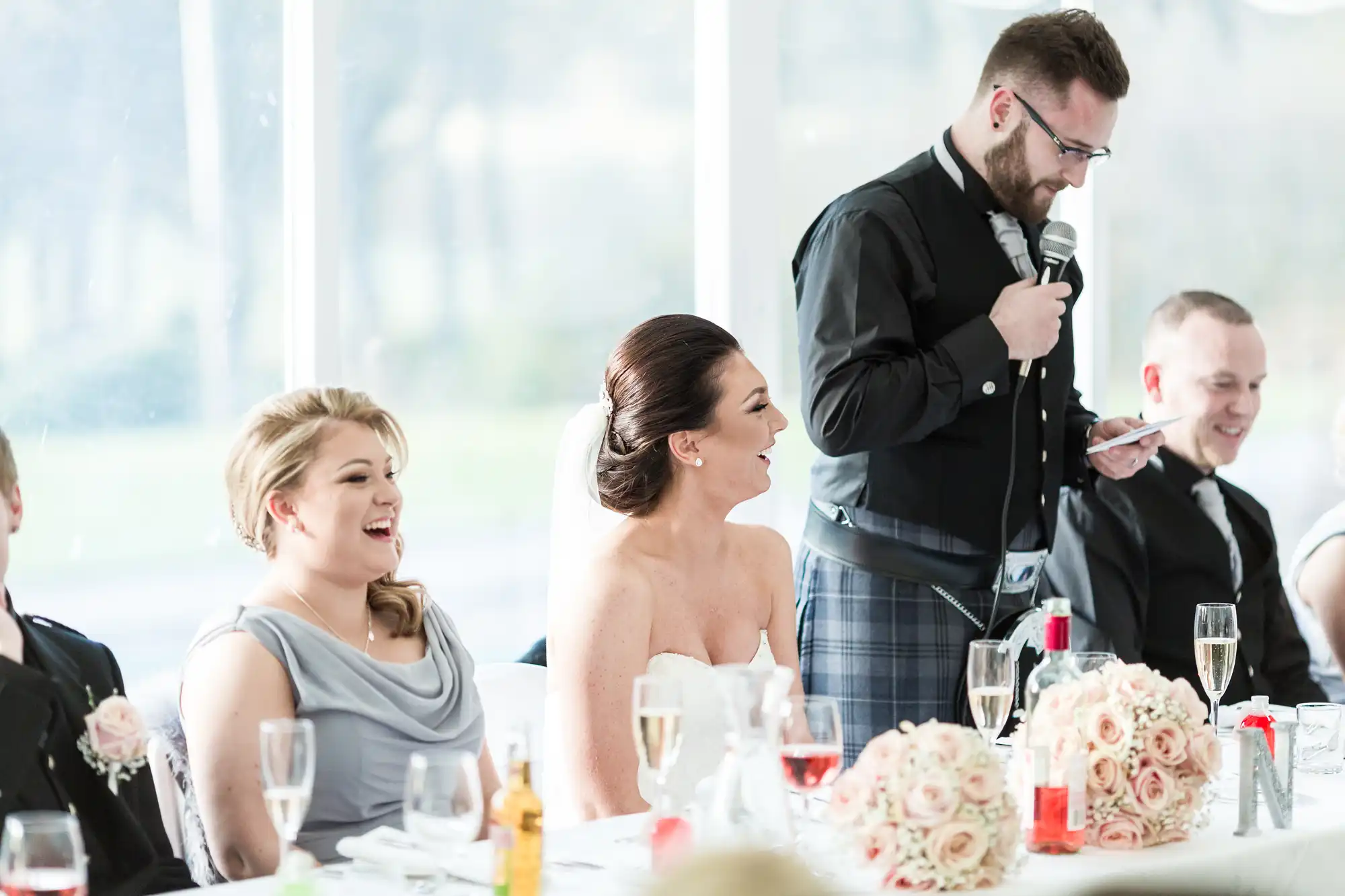 A man in a tartan kilt stands and speaks into a microphone at a wedding reception table. Two women in formal dresses and a man in a suit smile and listen. The table is decorated with flowers, wine, and champagne glasses. Bright daylight streams through large windows.