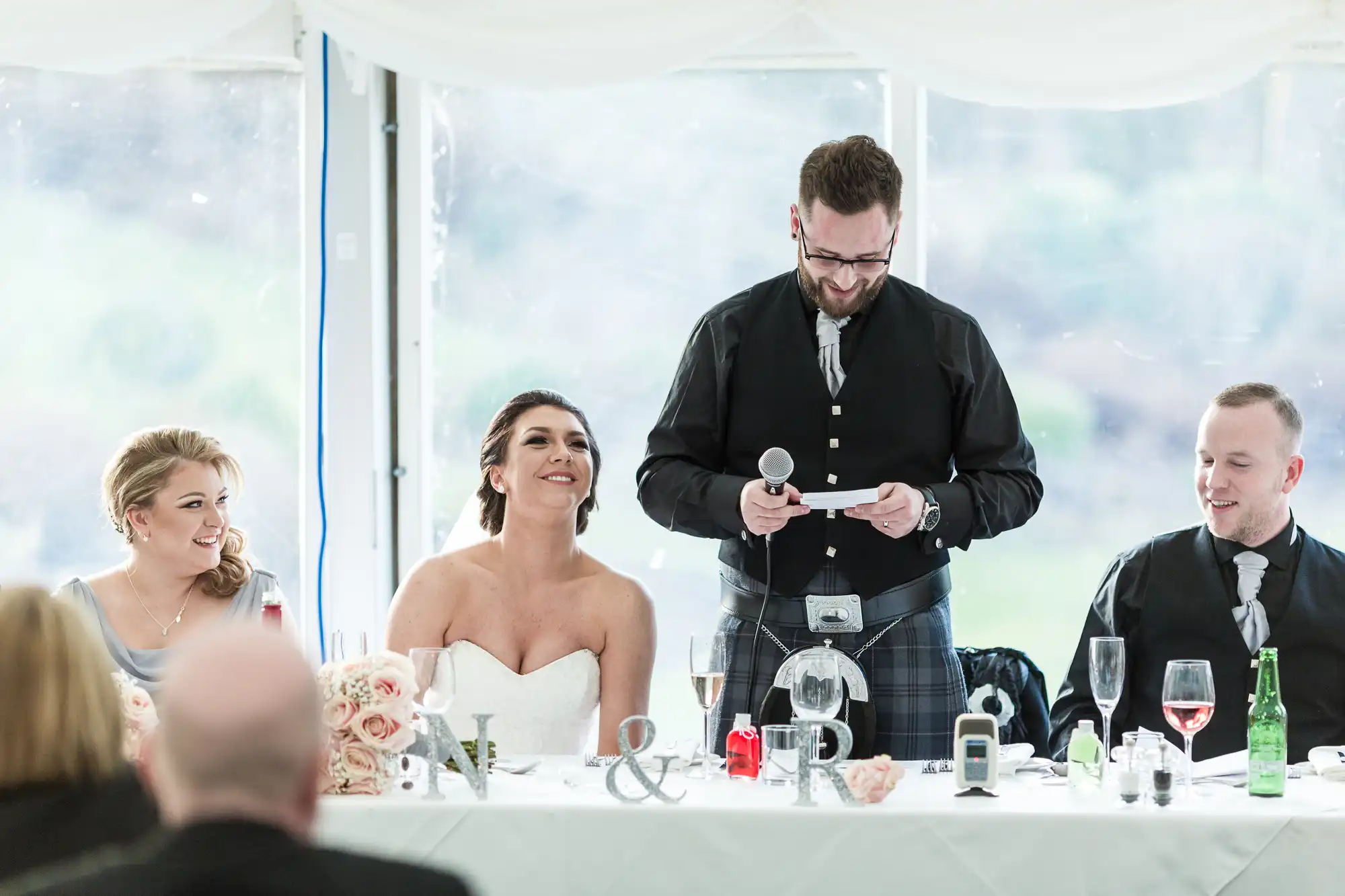 A bride in a strapless dress and three people sit at a decorated table while a man in a black jacket stands, smiling and reading from a card. Champagne glasses, flowers, and initials "N & R" decorate the white tablecloth. All appear happy and engaged.