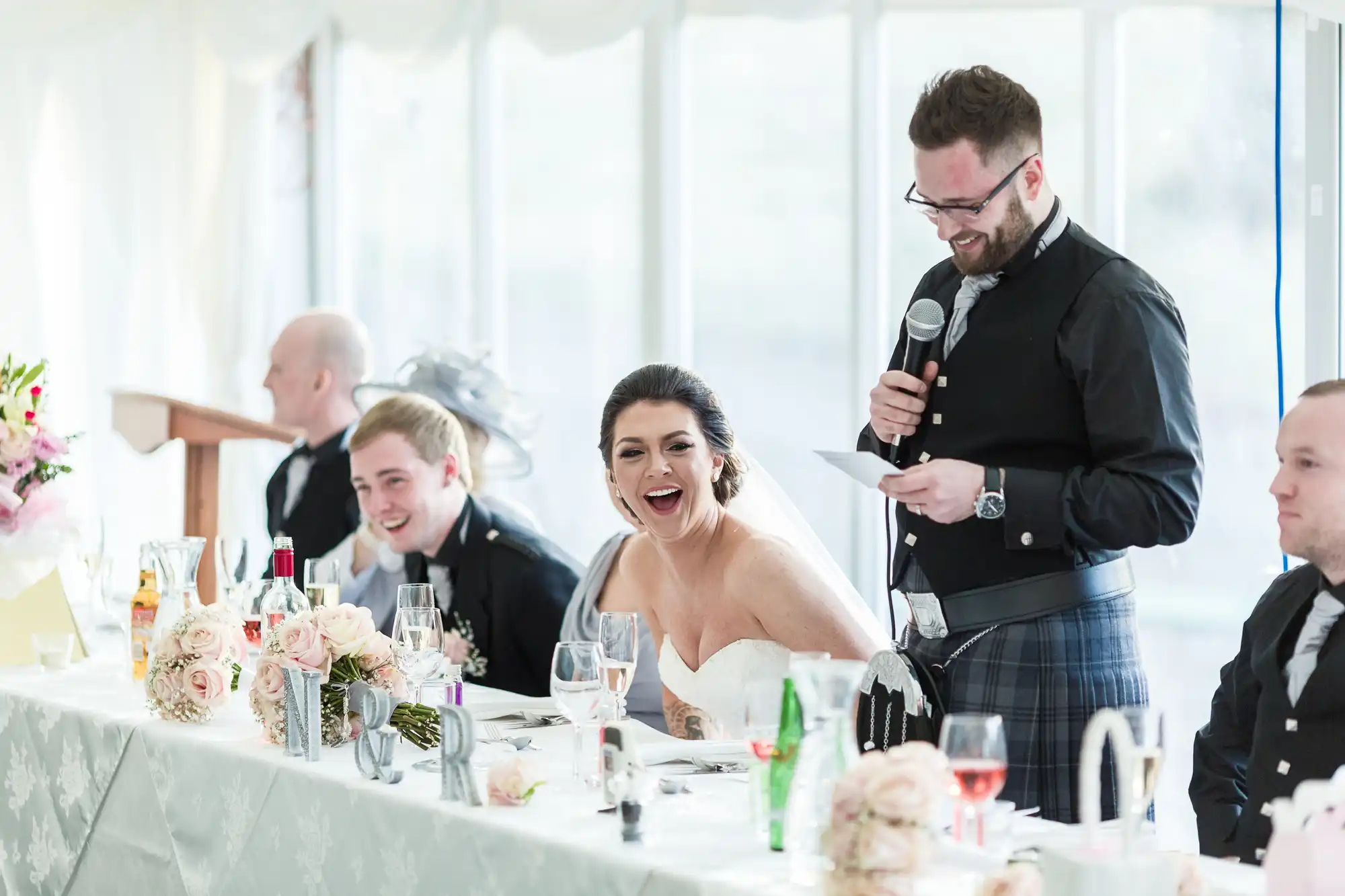 A bride in a strapless gown laughs joyfully at a long decorated table as a man in a kilt and jacket speaks into a microphone, holding a piece of paper. Other guests, dressed formally, smile and listen; the table is adorned with flowers, glasses, and elegant place settings.