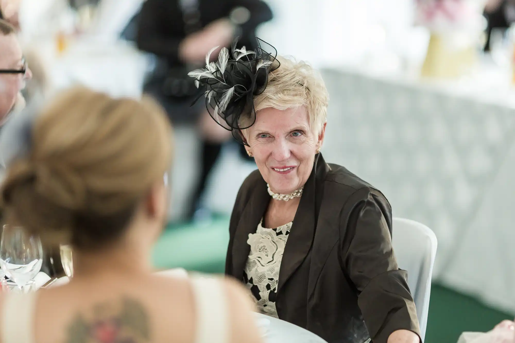 An older woman in a black jacket and floral dress, wearing a decorative black and white fascinator, smiles at the camera while seated at a table. A younger woman with a back tattoo and an updo sits in the foreground, slightly out of focus.